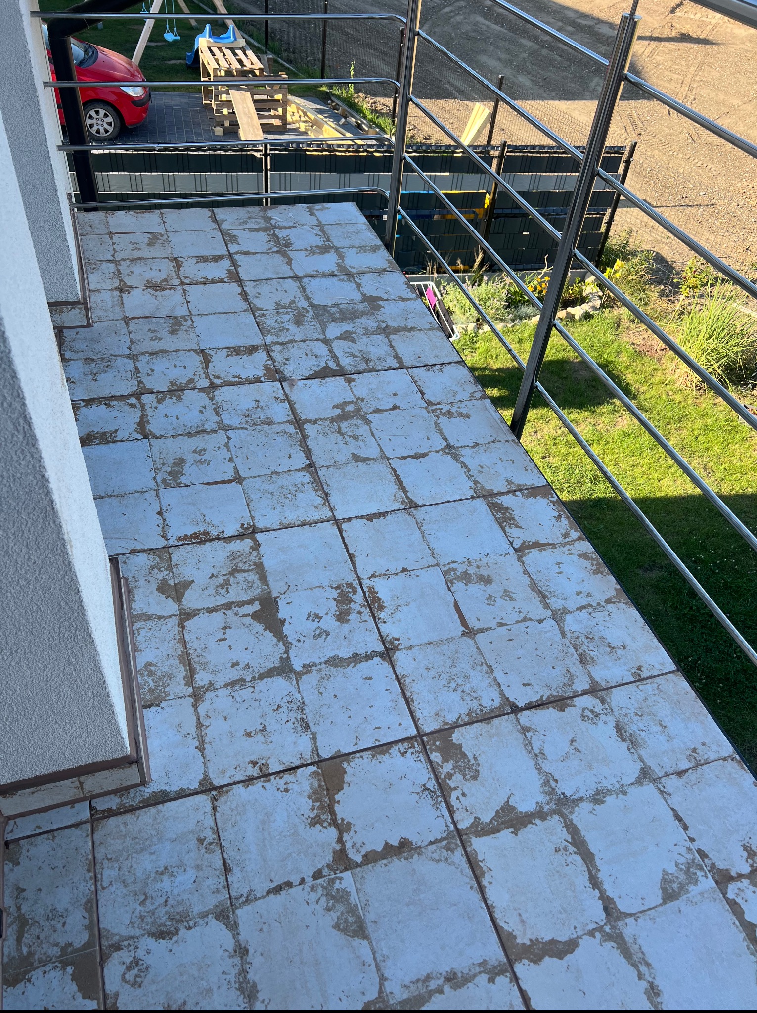 Balkon z płytkami wymagającymi renowacji; widoczne stare, popękane płytki w odcieniach błękitu i szarości, metalowa balustrada. W tle trawnik, plac zabaw i czerwony samochód.