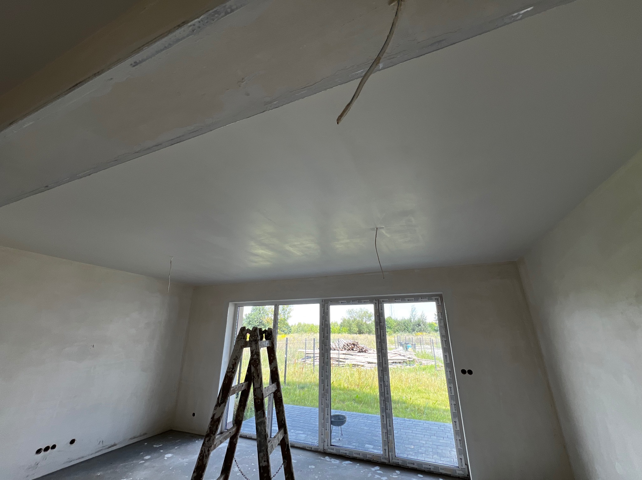 Wnętrze w trakcie prac wykończeniowych: gładzie na ścianach i suficie, drabina oparta o okno balkonowe, wystające kable elektryczne. Widok na zewnątrz.