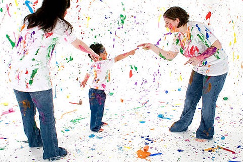 Dwie kobiety i dziecko w białych koszulkach i jeansach, ubrudzeni farbami, bawią się w białym pomieszczeniu z plamami farb, tworząc kolorowy chaos.