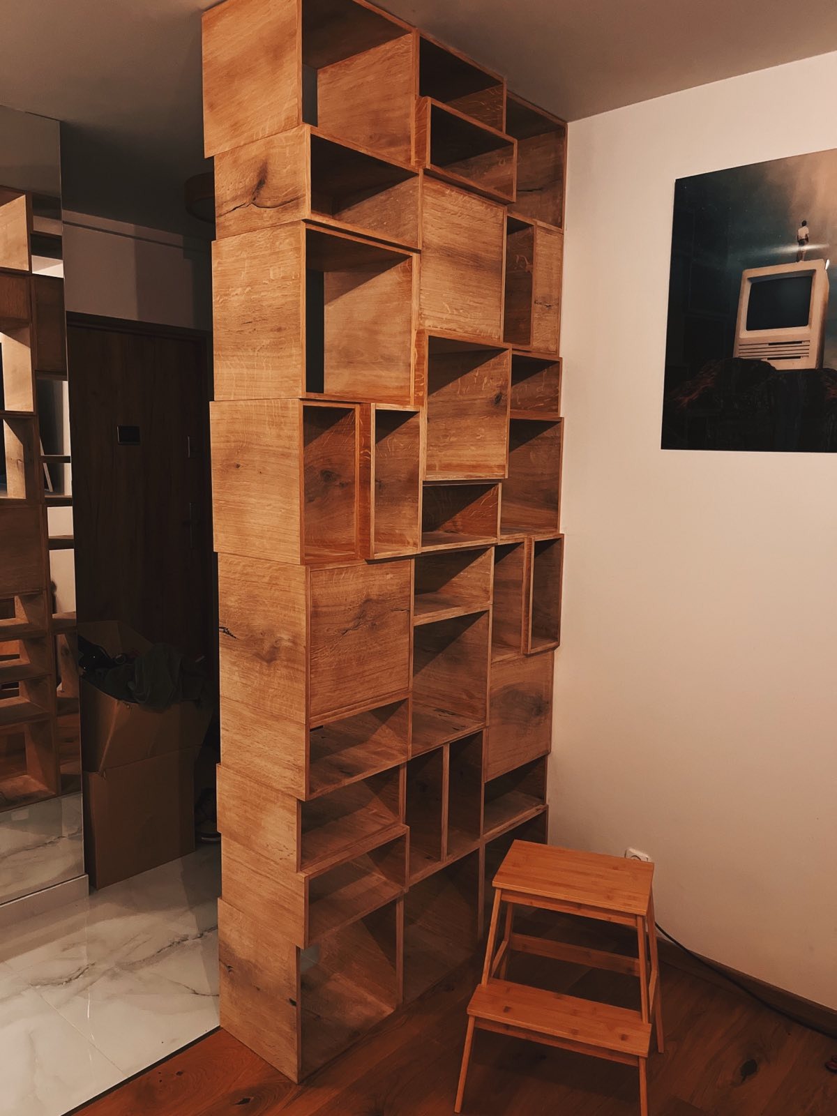 Drewniany regał modułowy, tworzący ściankę działową, z widoczną strukturą drewna i schodkami obok. Na ścianie plakat z motywem retro komputera.