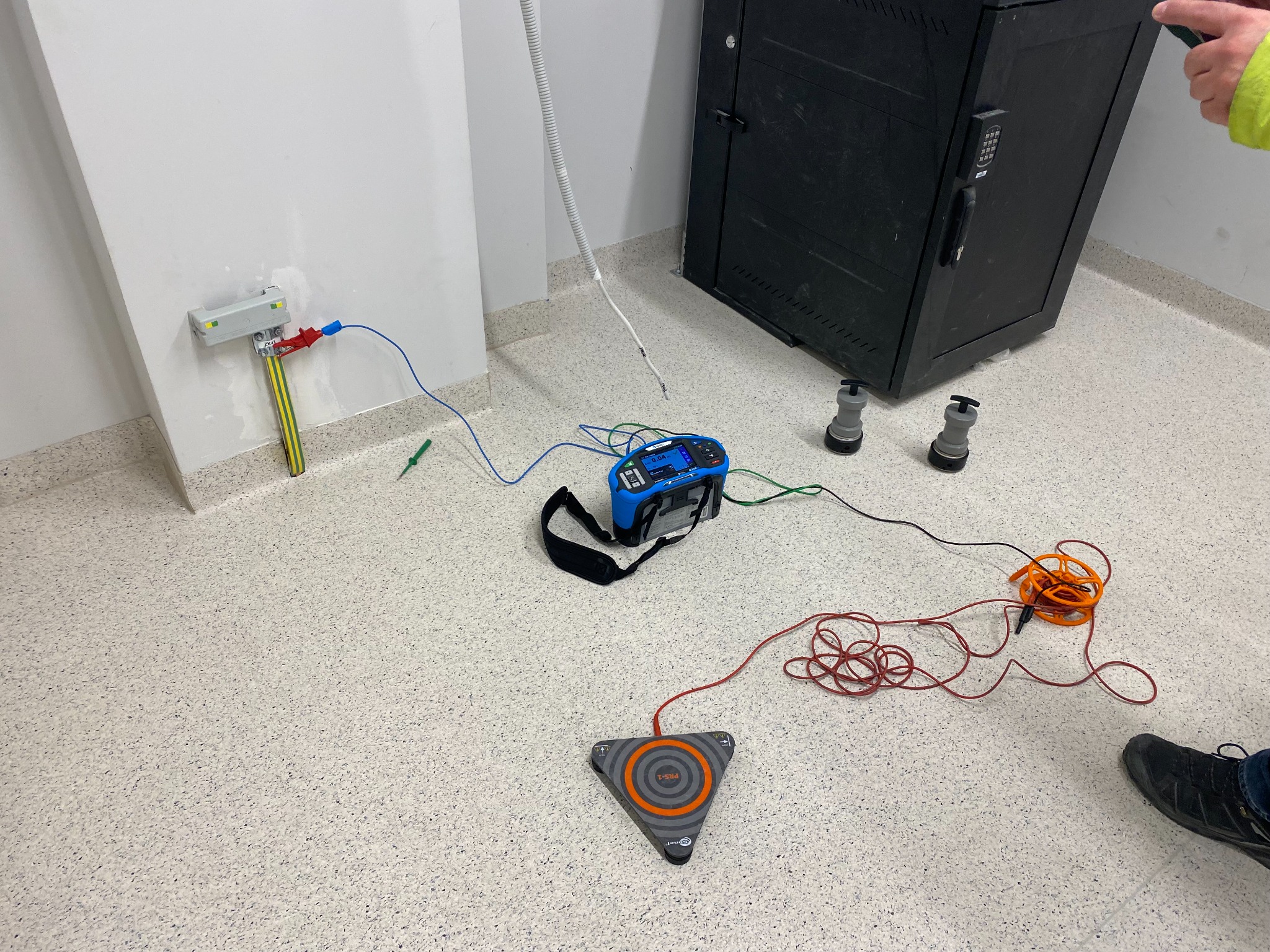 Podłączenie miernika do instalacji elektrycznej w pomieszczeniu z szafą rack. Widoczne przewody, miernik, uziemienie i fragment buta osoby wykonującej pomiary.