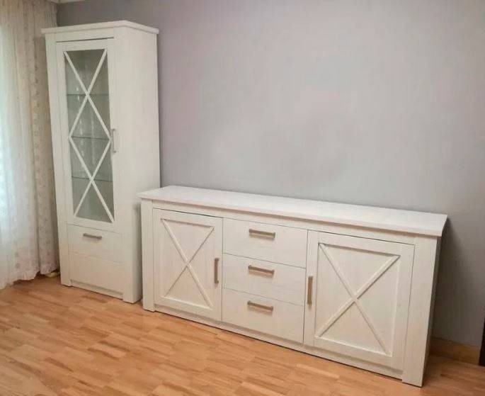 Biały kredens i witryna z przeszkleniami, stylizowane na rustykalne, z drewnianymi krzyżami na frontach, na parkiecie w jasnym odcieniu.