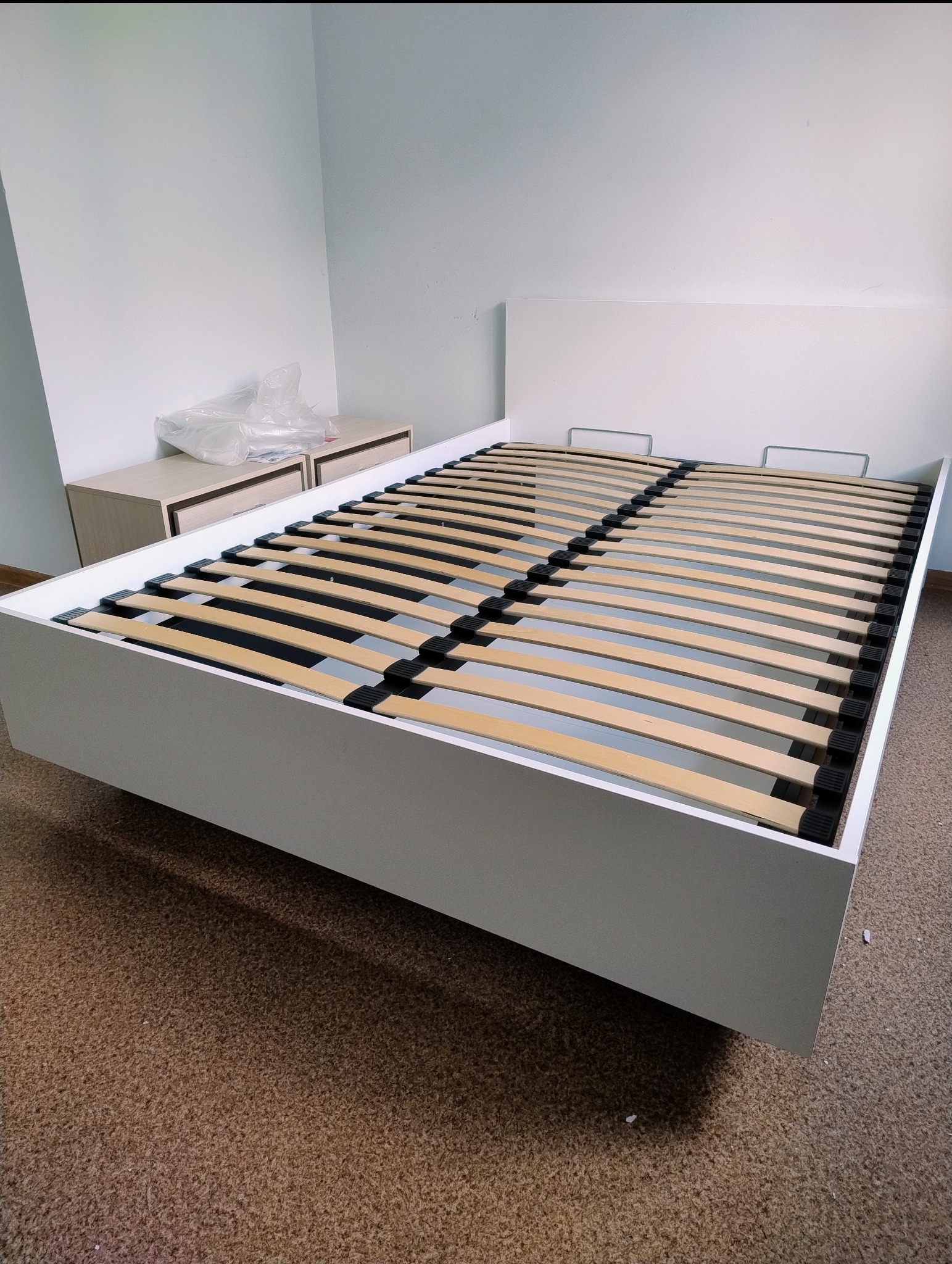 Białe łóżko z drewnianym stelażem i zagłówkiem, z szafkami nocnymi w tle. Widok z dołu, ukazujący konstrukcję mebla po złożeniu.