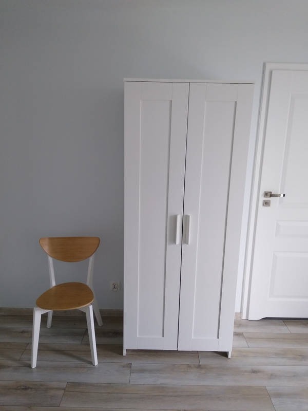 Biała szafa dwudrzwiowa z uchwytami, obok drewniane krzesło na tle szarej ściany i białych drzwi. Podłoga z paneli imitujących drewno.