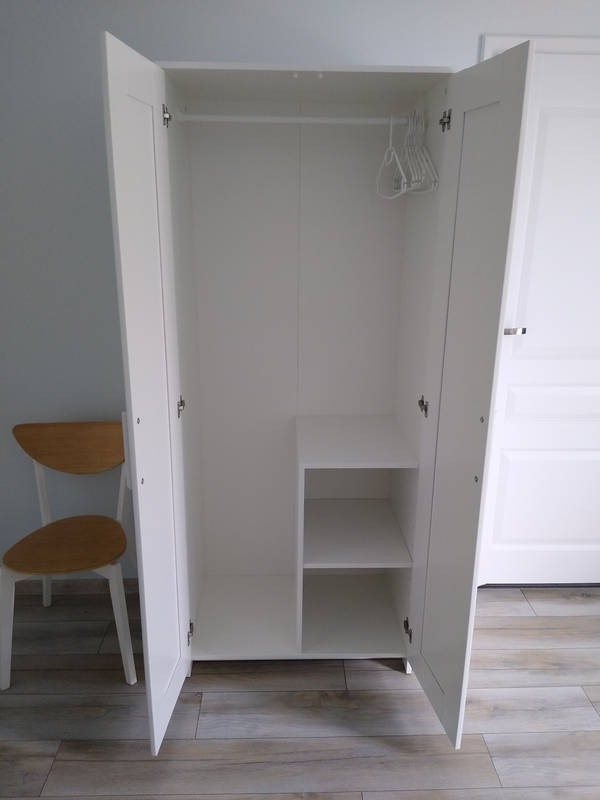 Biała szafa z otwartymi drzwiami, półkami i wieszakami, stojąca na drewnianej podłodze. Obok krzesło. Widok wnętrza szafy.