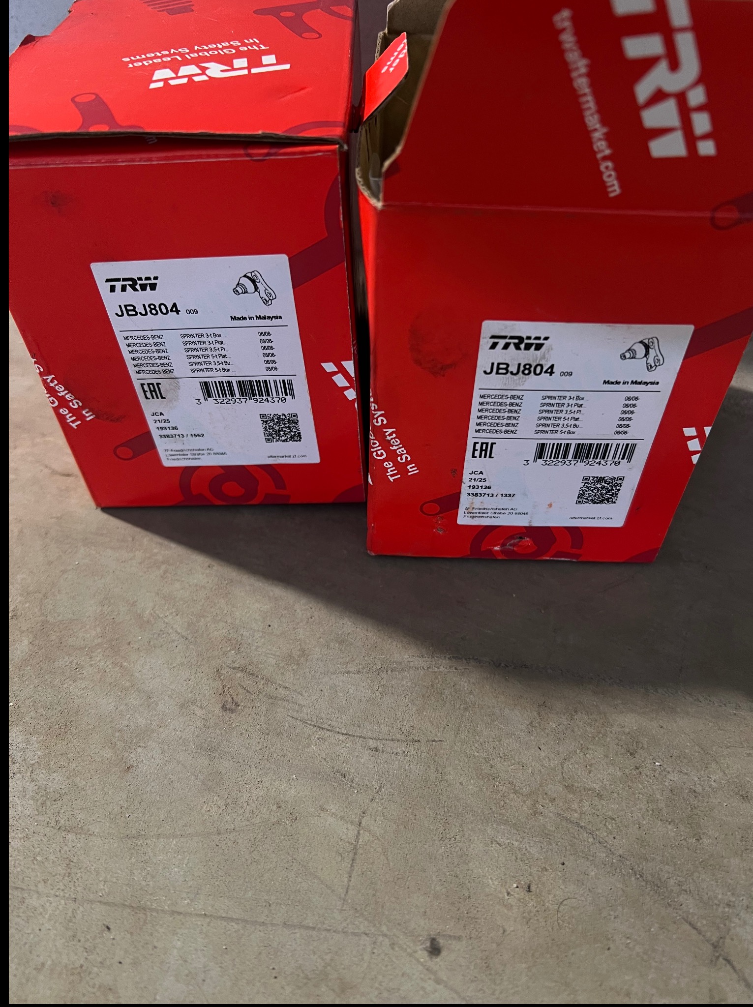 Dwa czerwone pudełka TRW JBJ804 z częściami zamiennymi do Mercedesa Sprintera, widoczne etykiety z informacjami o modelu i dacie produkcji, na szarym tle podłogi.