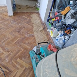 Jarosław Bereza - Parkiet w jodełkę po cyklinowaniu, widoczna różnica w kolorze i strukturze drewna. W tle narzędzia i materiały budowlane. Maszyna do cyklinowania na pierwszym planie.