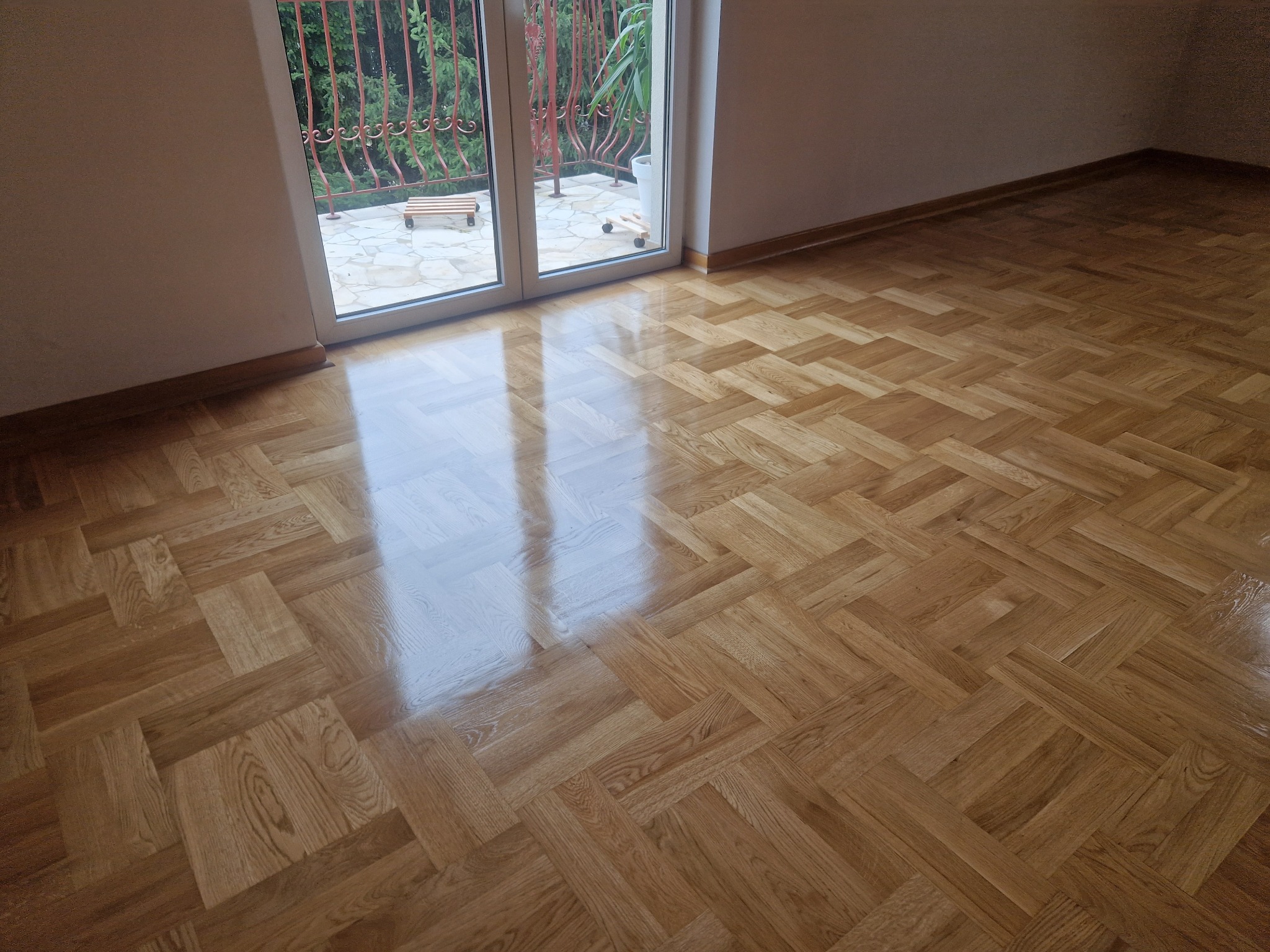 Błyszcząca, odnowiona drewniana podłoga parkietowa w jodełkę, z widocznym wyjściem na balkon i fragmentem zieleni w tle. Świeżo po renowacji, bez śladów użytkowania.