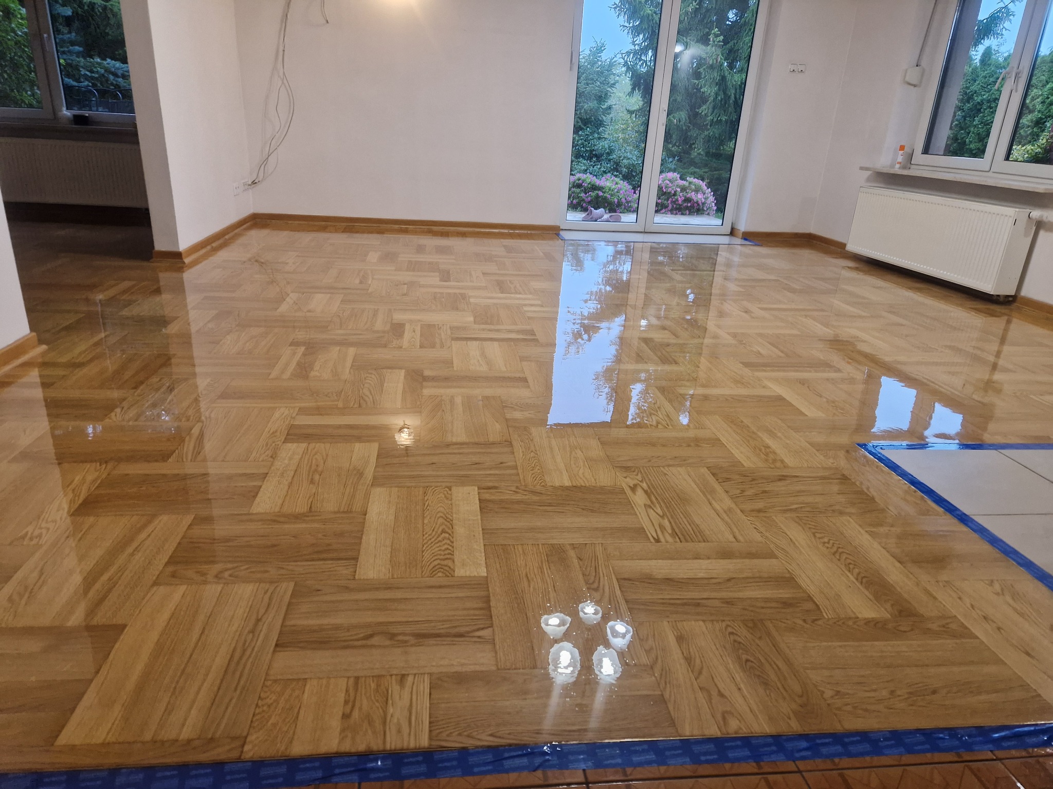 Błyszcząca, odnowiona parkietowa podłoga w jodełkę po renowacji, zabezpieczona folią malarską przy ścianach, z widocznymi odbiciami światła.