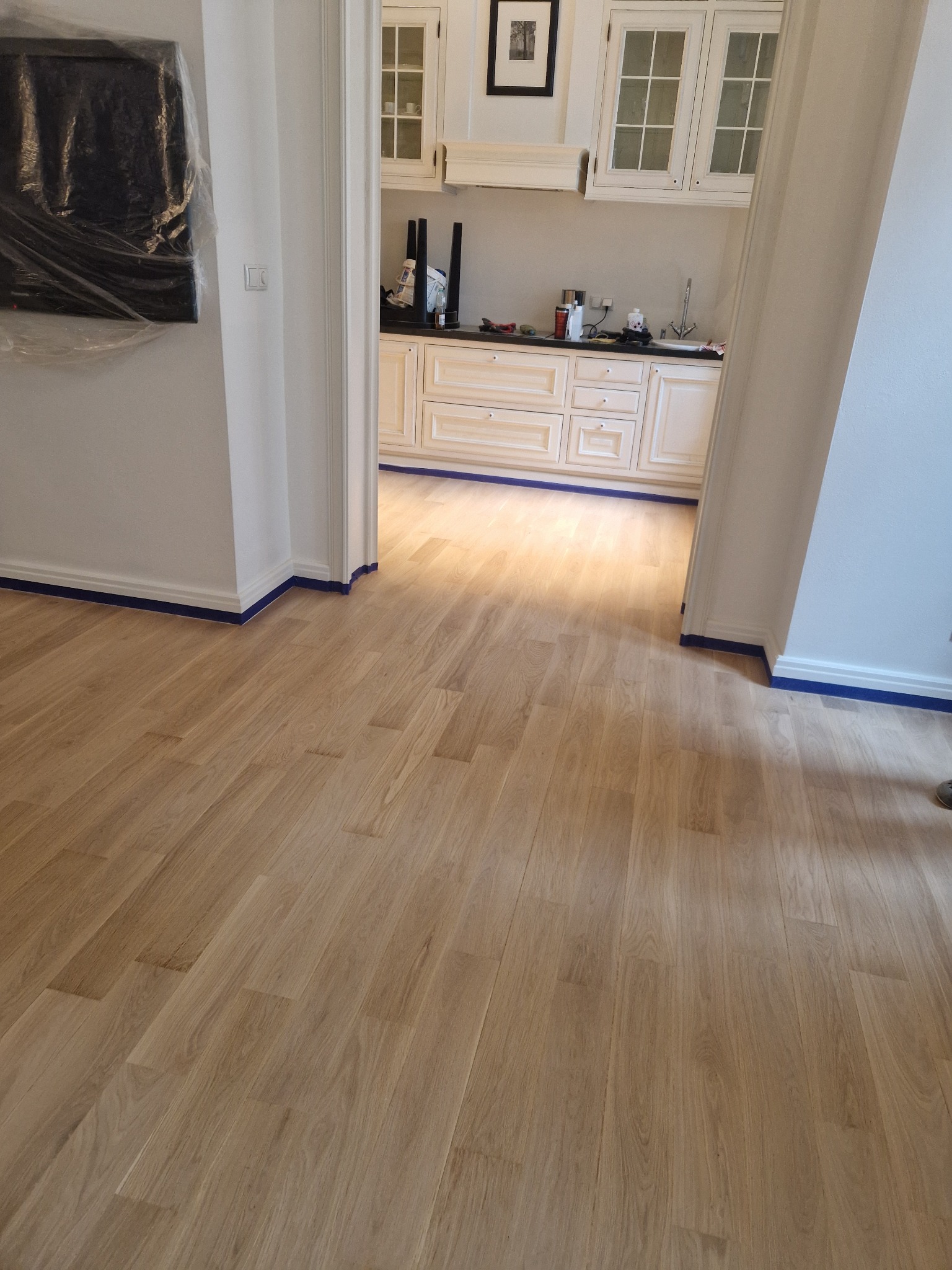 Jasna, świeżo wycyklinowana podłoga parkietowa z zabezpieczoną listwą przypodłogową niebieską taśmą malarską, widok na kuchnię w tle.
