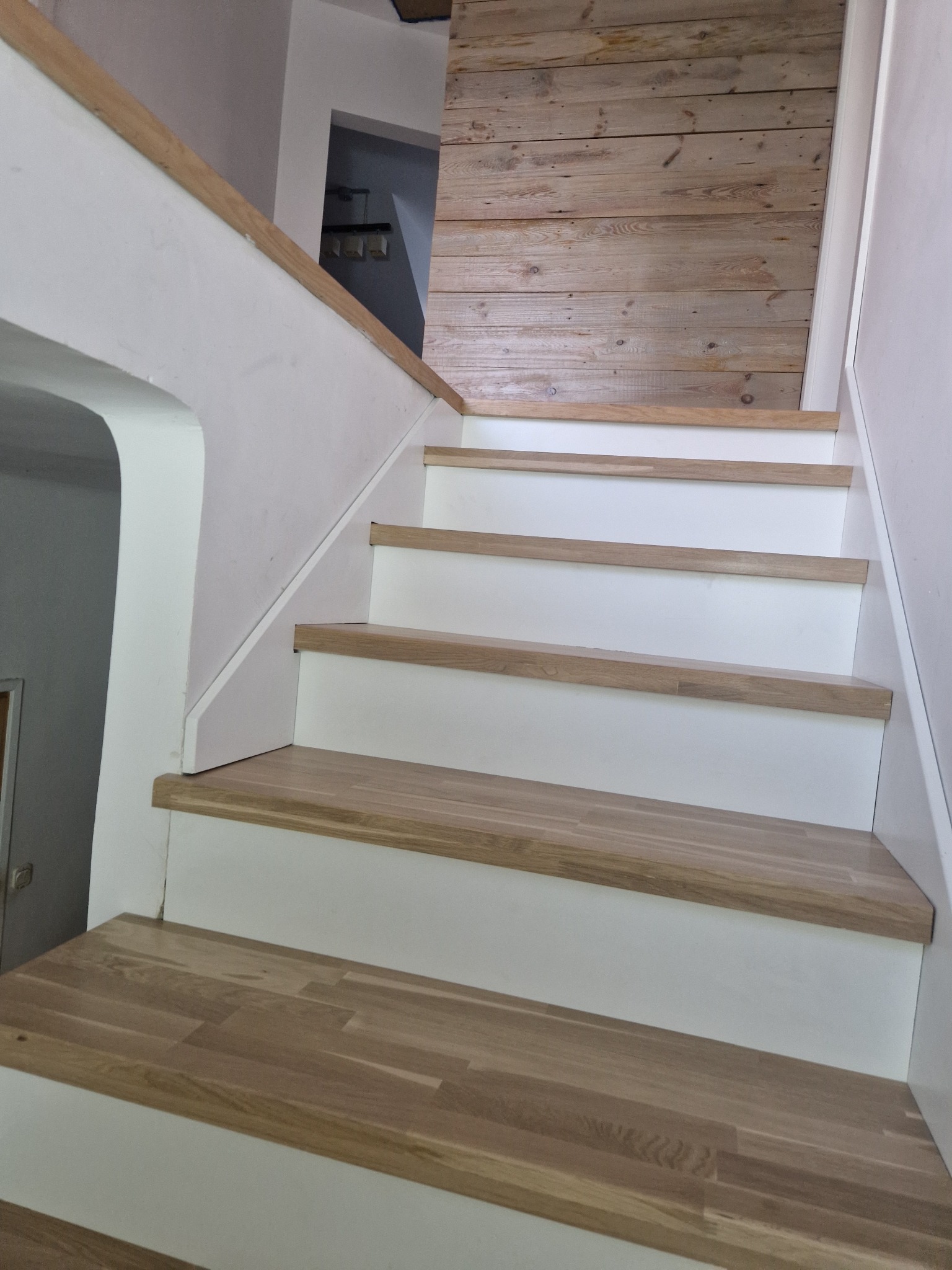 Odnowione schody z drewnianymi stopniami i białymi podstopnicami, widok z dołu, z fragmentem drewnianej ściany w tle. Schody po renowacji.
