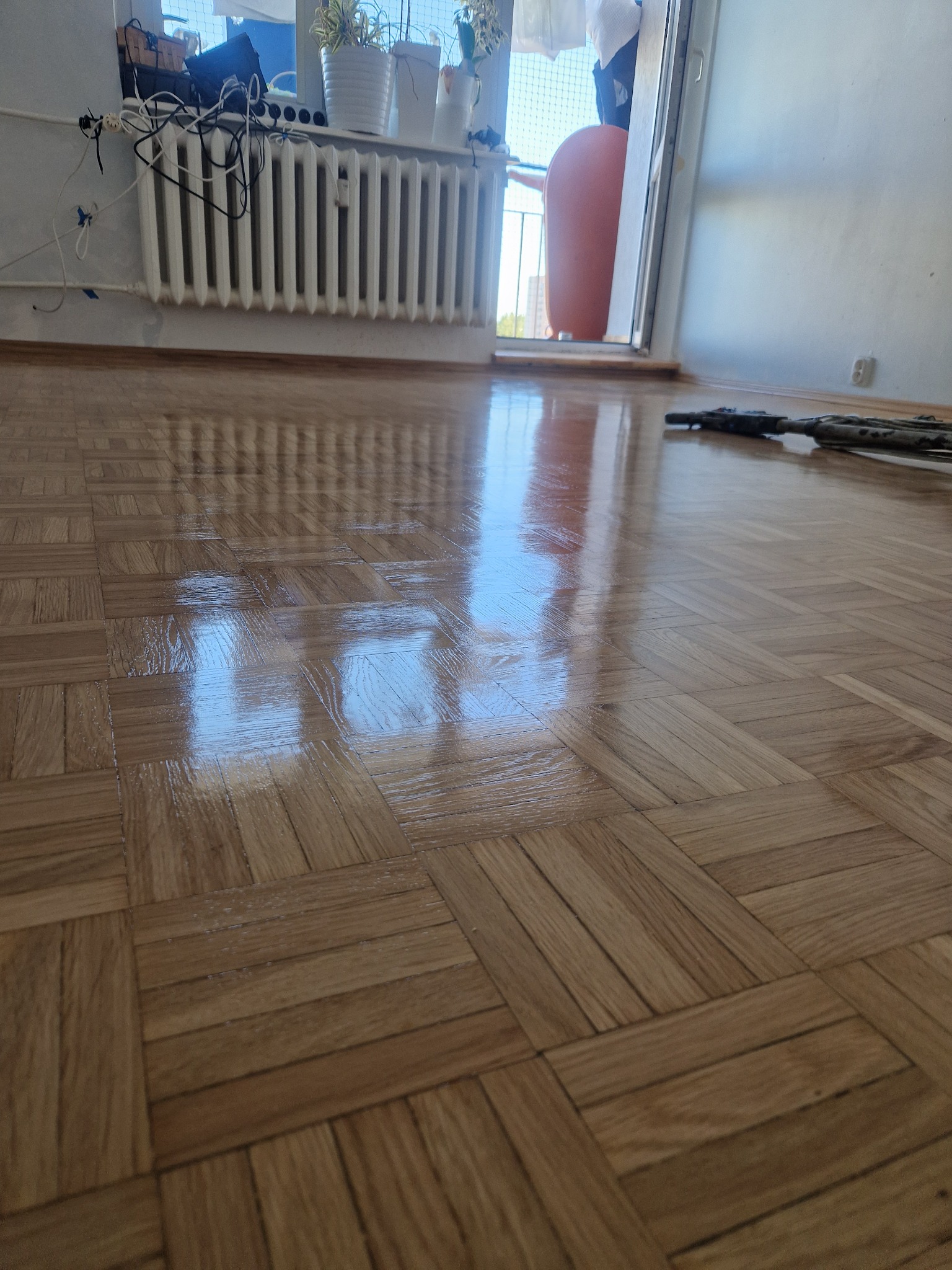Błyszcząca, odnowiona podłoga parkietowa w jodełkę, odbijająca światło okna. Widoczne narzędzia do polerowania pod ścianą, kaloryfer i rośliny w tle.