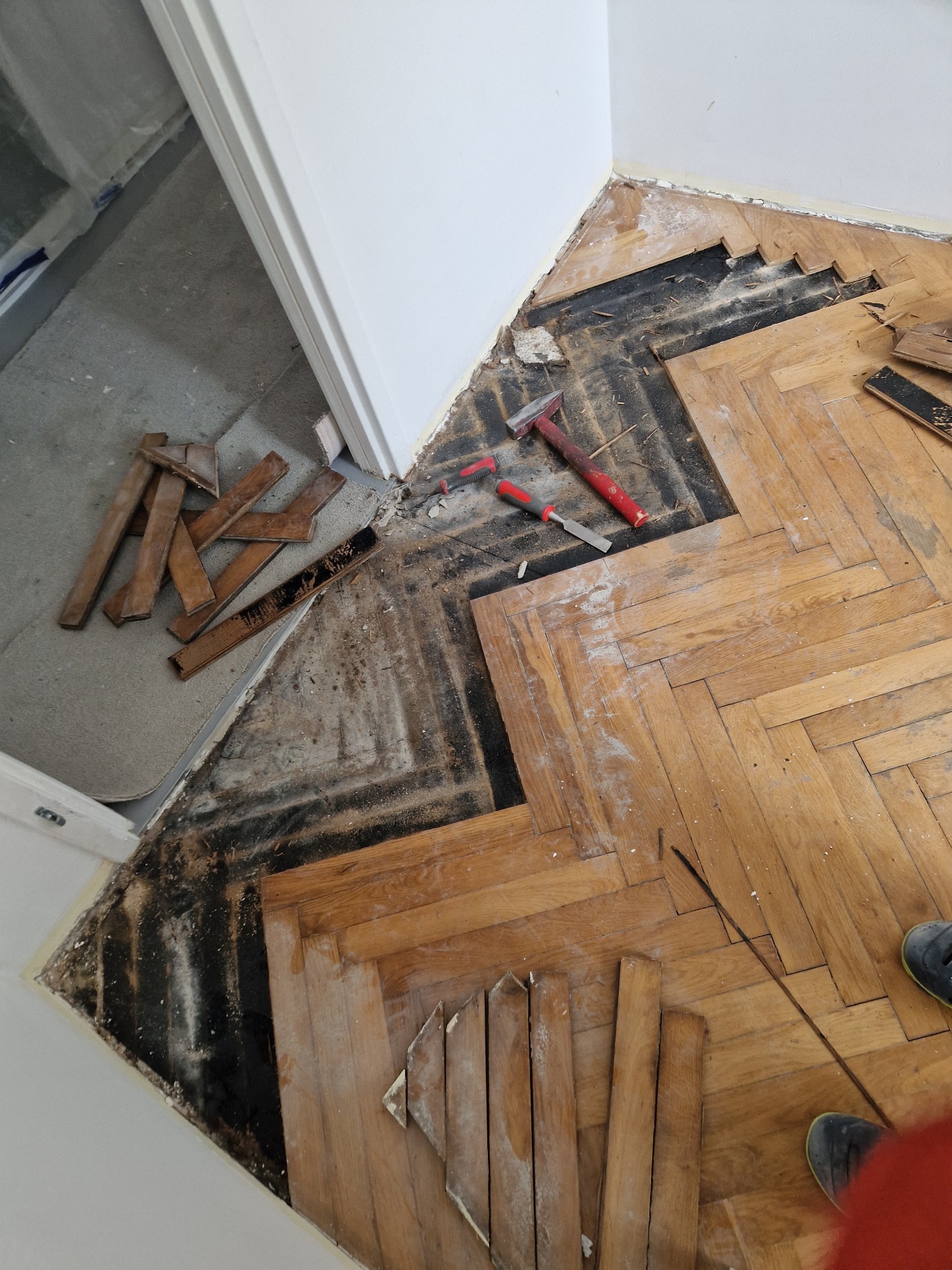 Parkiet w jodełkę w trakcie cyklinowania, z widocznymi fragmentami starej, ciemnej warstwy i narzędziami: młotkiem i dłutem. Część desek usunięta.