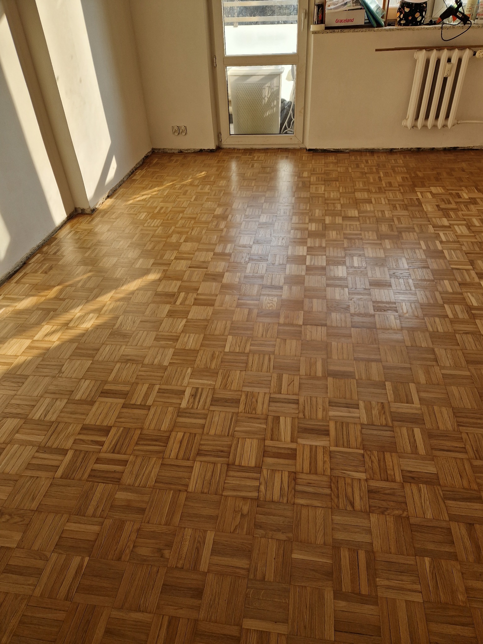 Parkiet ułożony w kwadraty, po renowacji, lśniący w promieniach słońca w Ząbkach. Widoczne ślady po listwach przypodłogowych, grzejnik i okno.