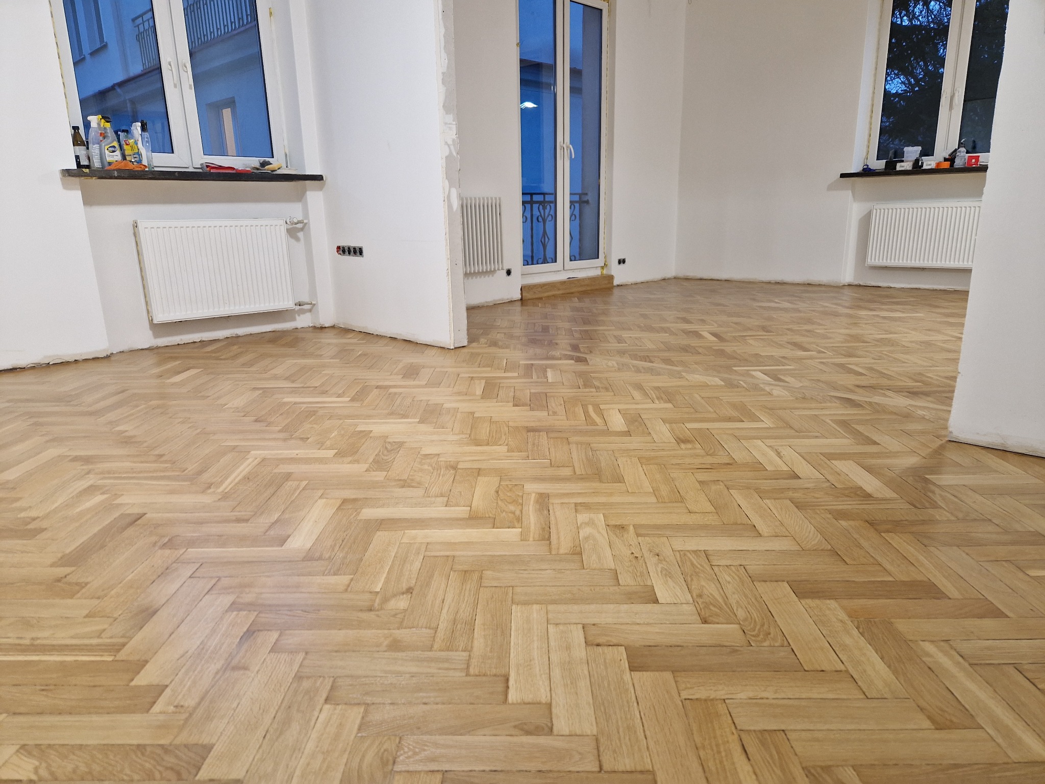 Jasny parkiet ułożony w jodełkę w odnowionym pokoju z białymi ścianami i grzejnikami. Widoczne okna i drzwi balkonowe. Podłoga po renowacji.