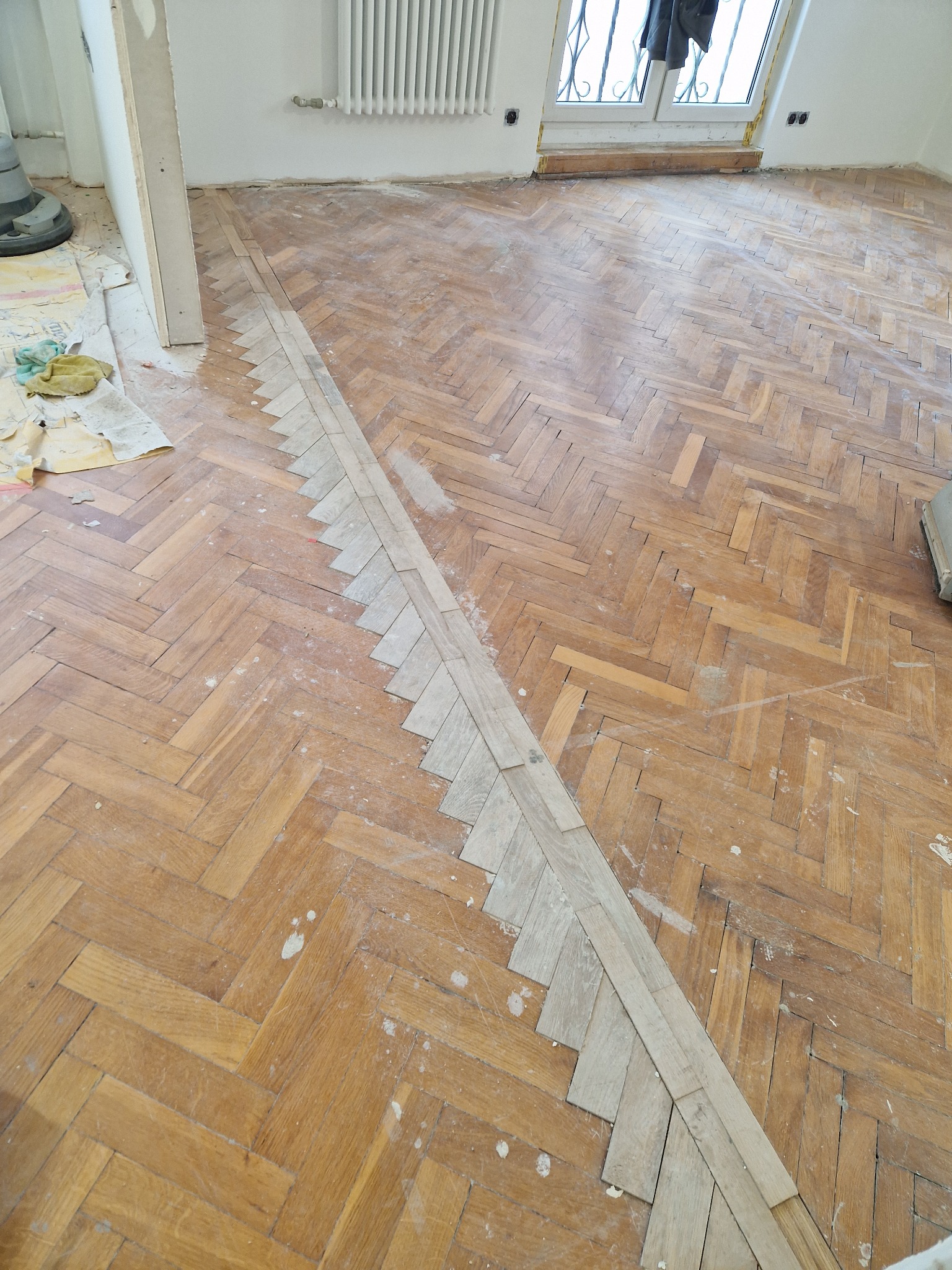 Parkiet w jodełkę w trakcie renowacji: częściowo przeszlifowany, z widocznymi fragmentami nowej, jaśniejszej klepki. Fragment maszyny do cyklinowania w tle.