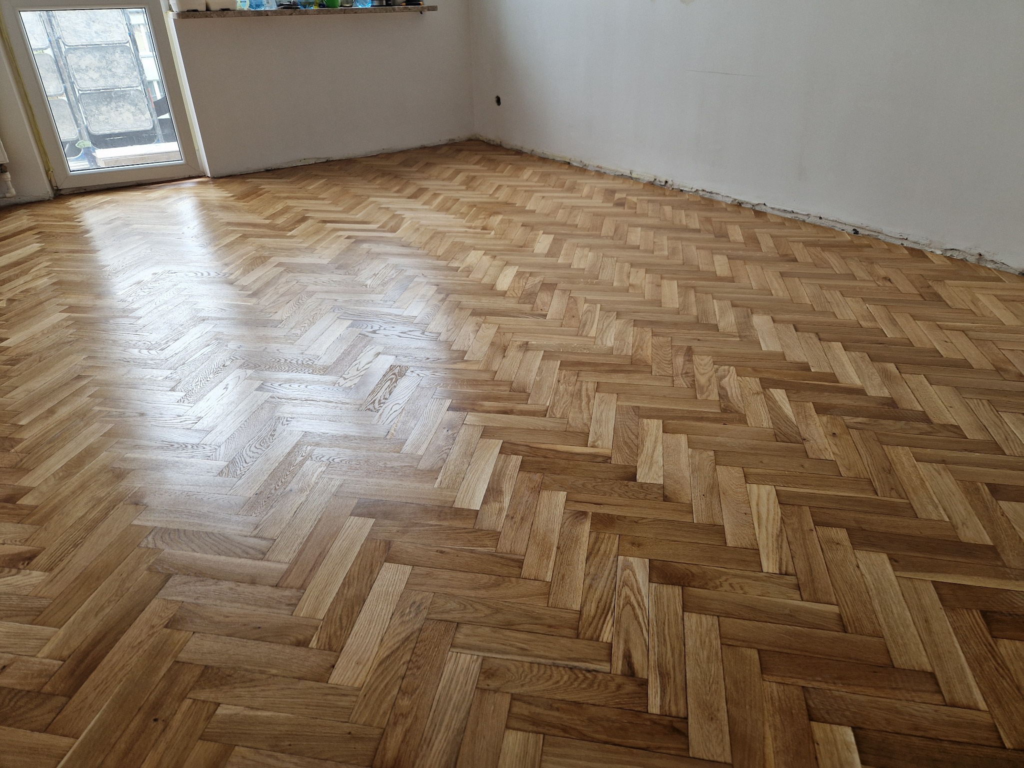 Parkiet ułożony w jodełkę po renowacji, widoczne ślady po szlifowaniu przy ścianach, naturalne światło z okna podkreśla strukturę drewna.