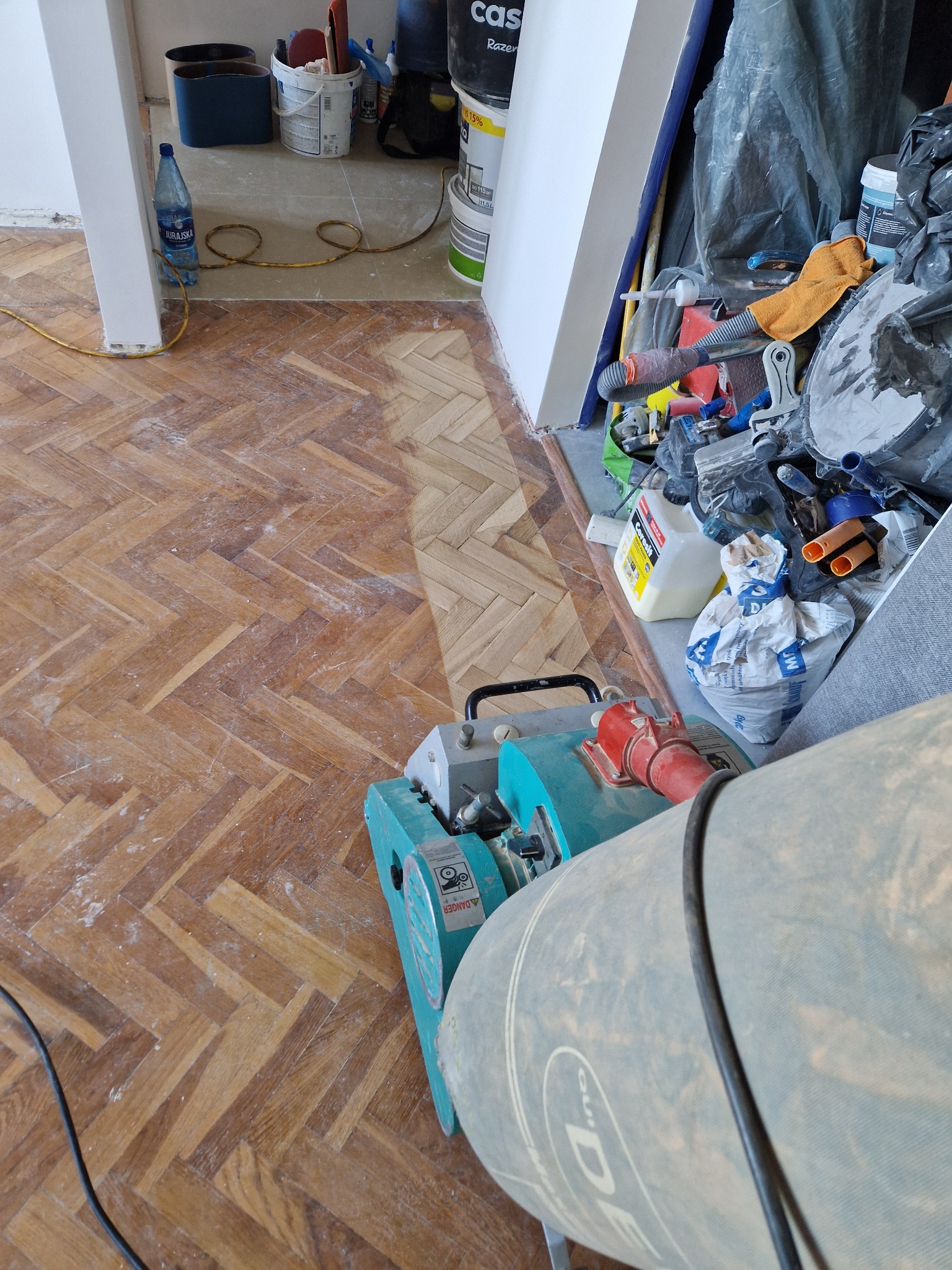 Parkiet w jodełkę po cyklinowaniu, widoczna różnica w kolorze i strukturze drewna. W tle narzędzia i materiały budowlane. Maszyna do cyklinowania na pierwszym planie.