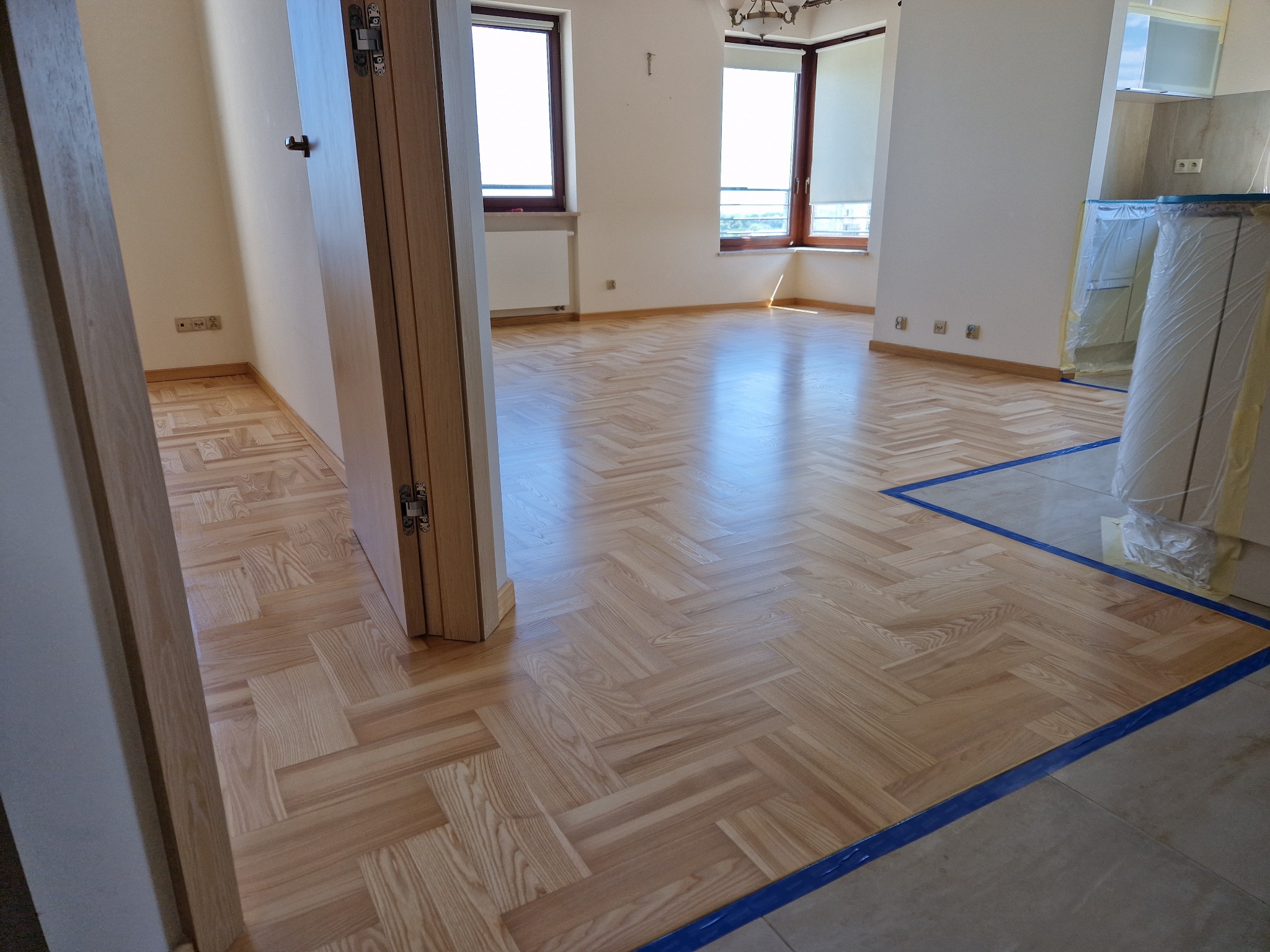 Parkiet ułożony w jodełkę, po renowacji, z widocznym zabezpieczeniem folią malarską przy łączeniu z płytkami. Jasne, przestronne wnętrze z oknami.