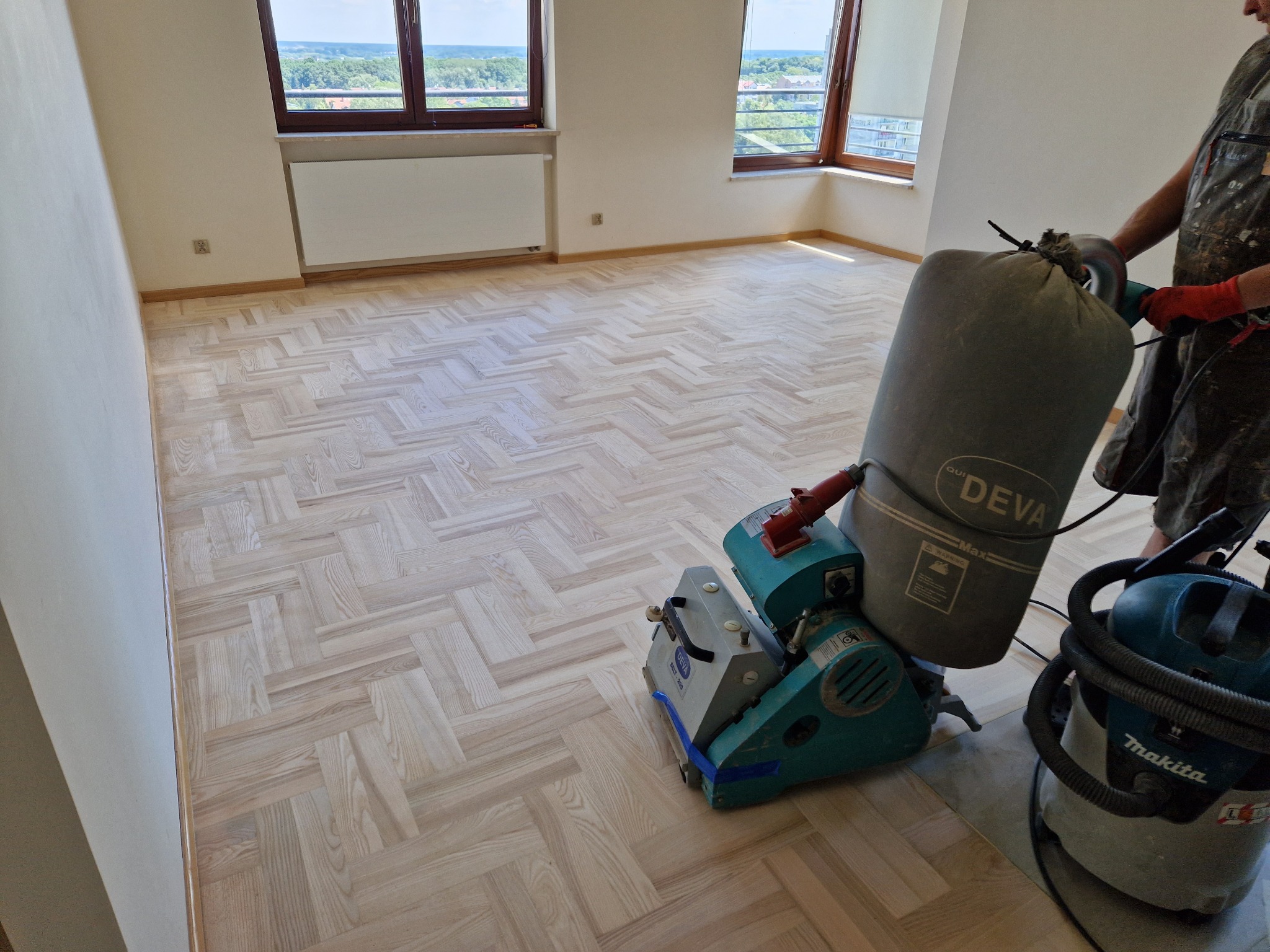 Parkiet ułożony w jodełkę po zabiegach renowacyjnych. Widoczna maszyna do cyklinowania Deva i odkurzacz Makita. Jasne wnętrze z dużymi oknami i widokiem na zieleń.