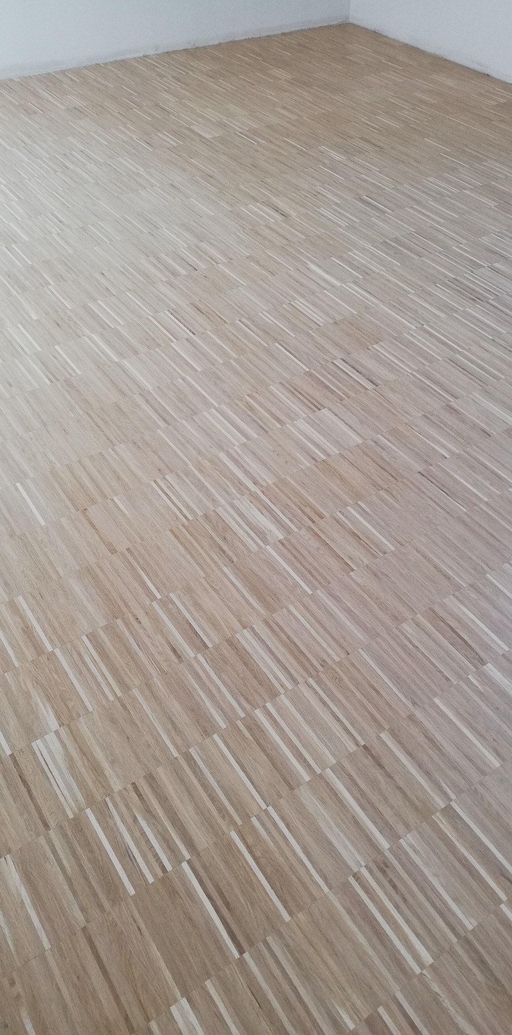 Ukończona podłoga parkietowa w jasnych odcieniach drewna, ułożona w geometryczny wzór. Widok na fragment pomieszczenia z białymi ścianami.