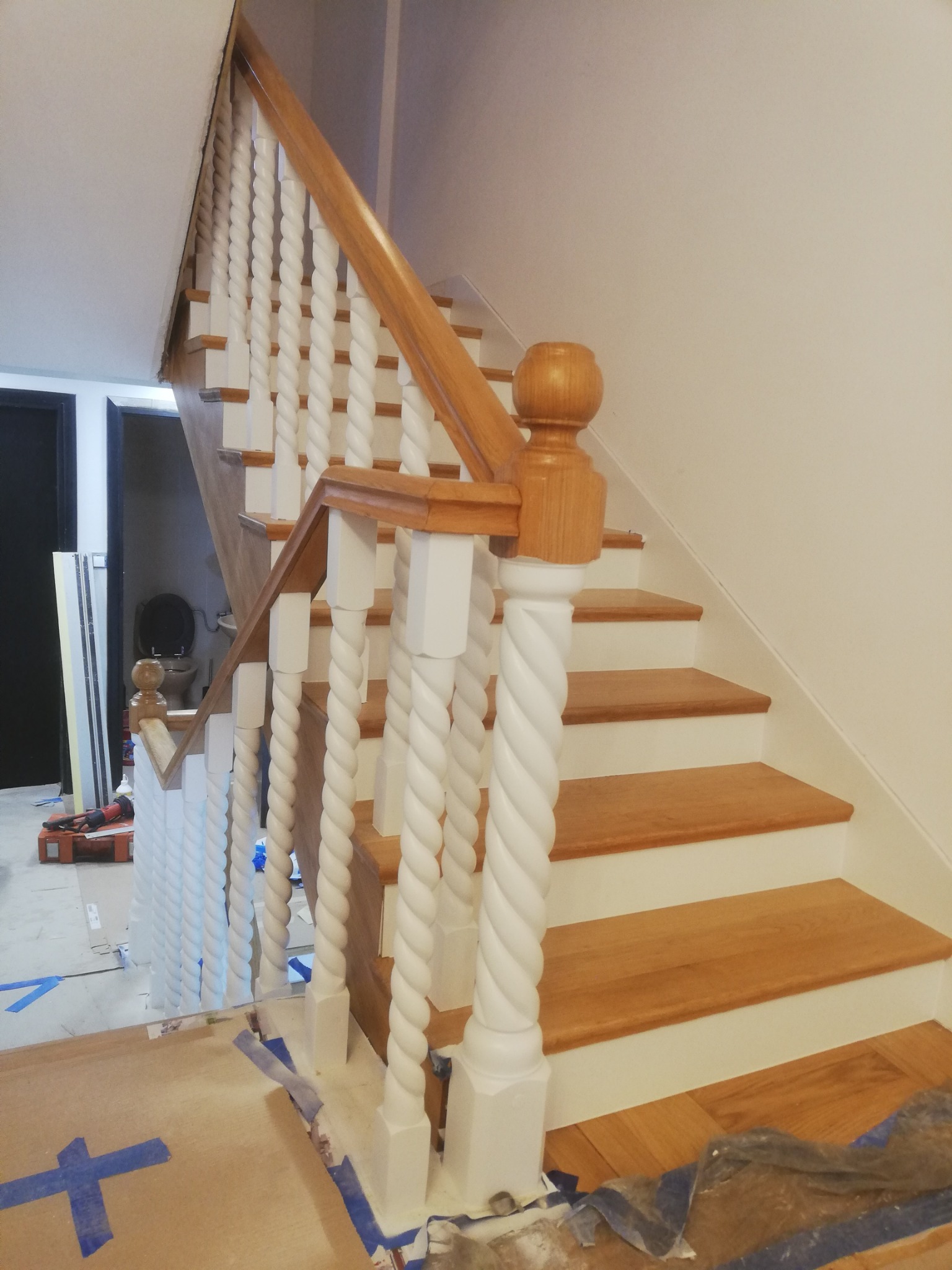 Odnowione schody drewniane z białymi balustradami o spiralnym wzorze, widoczne ślady prac wykończeniowych, jasne oświetlenie, perspektywa od dołu.