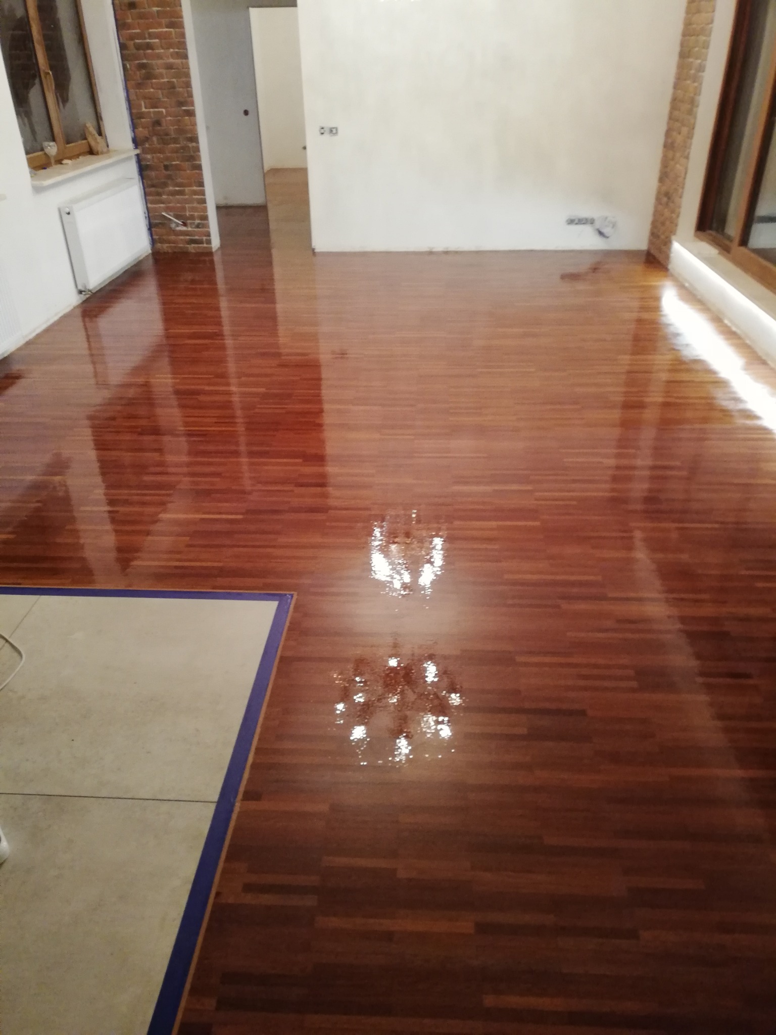 Błyszcząca, odnowiona podłoga parkietowa z ciemnego drewna, po zabiegu lakierowania, odbijająca światło lampy. Widoczne fragmenty ścian z cegły i gładzi.