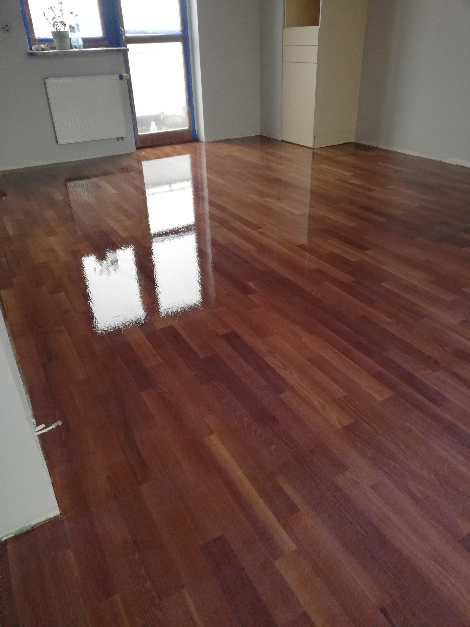 Błyszcząca, świeżo polakierowana parkietowa podłoga w odcieniach brązu odbija światło okna. Widoczne ślady po taśmie malarskiej przy ścianie.