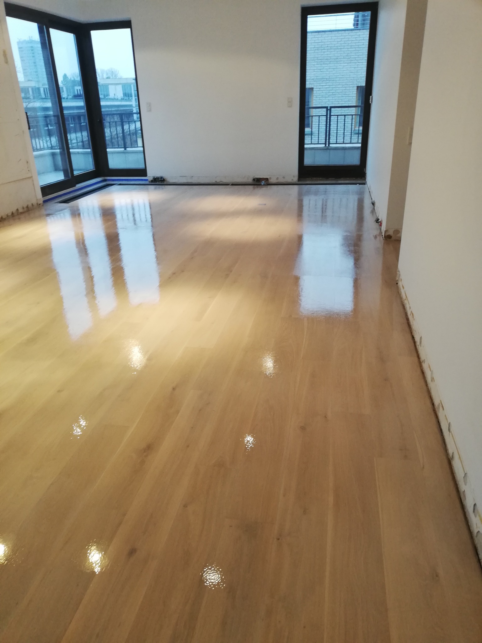 Błyszcząca, polakierowana podłoga parkietowa w pustym pokoju z oknami balkonowymi. Widoczne ślady po listwach przypodłogowych.