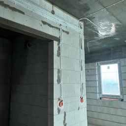 Bartosz Pękala usługi Elektryczne. Usługi Ogrodnicze - Białe rurki instalacji elektrycznej prowadzone po ścianach z betonu komórkowego w surowym wnętrzu. Widoczne puszki elektryczne i okno z folią ochronną.