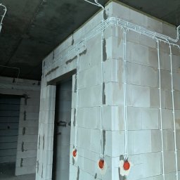 Bartosz Pękala usługi Elektryczne. Usługi Ogrodnicze - Białe kable elektryczne prowadzone po ścianach z betonu komórkowego, widoczne puszki instalacyjne. Surowy stan wykończenia wnętrza.