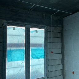 Bartosz Pękala usługi Elektryczne. Usługi Ogrodnicze - Białe przewody elektryczne prowadzone po suficie i ścianie w surowym wnętrzu z oknem zabezpieczonym folią. Widoczne puszki instalacyjne.