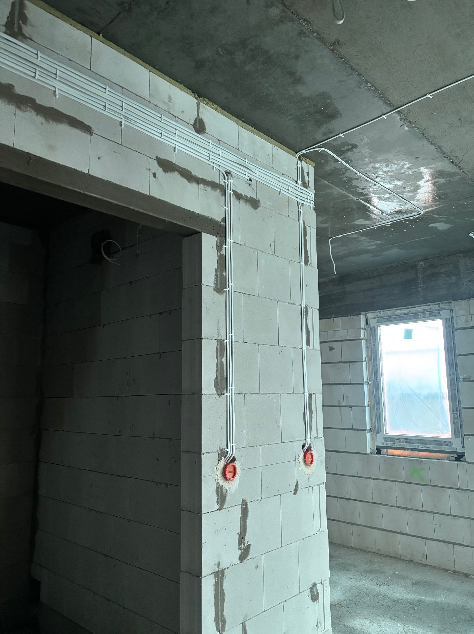 Białe rurki instalacji elektrycznej prowadzone po ścianach z betonu komórkowego w surowym wnętrzu. Widoczne puszki elektryczne i okno z folią ochronną.