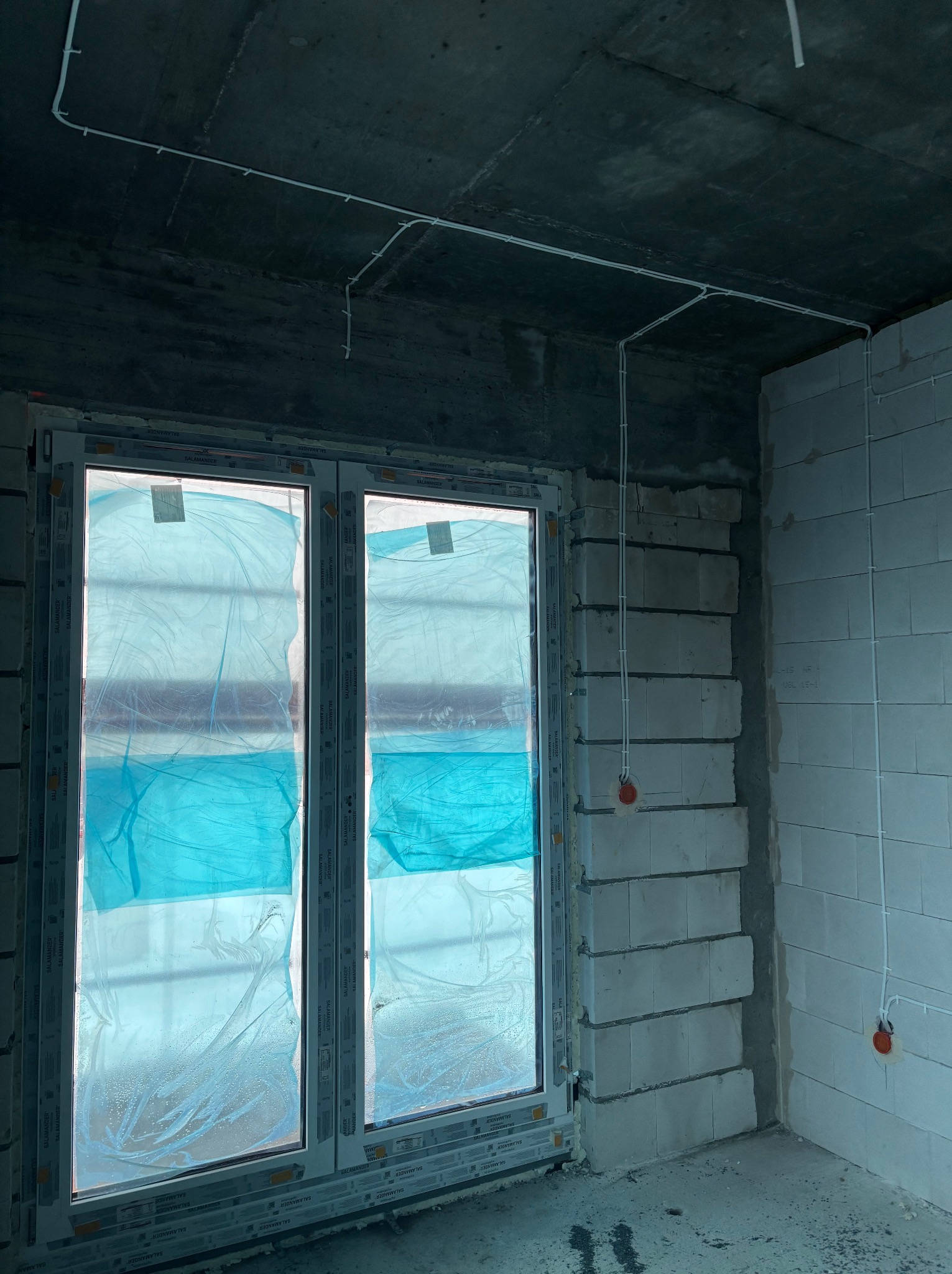 Białe przewody elektryczne prowadzone po suficie i ścianie w surowym wnętrzu z oknem zabezpieczonym folią. Widoczne puszki instalacyjne.