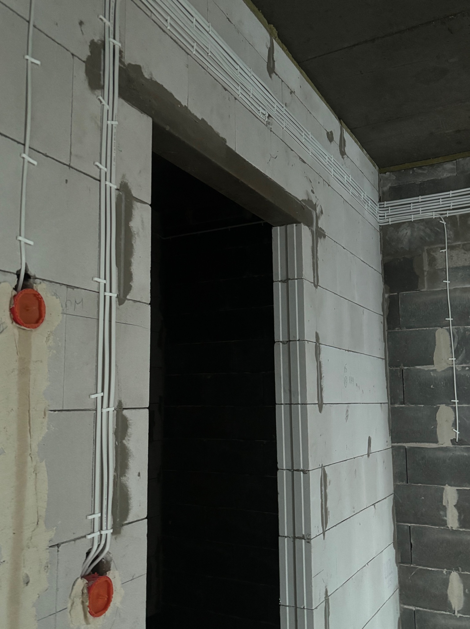 Białe kable elektryczne ułożone wzdłuż surowej ściany z betonu komórkowego, widoczne puszki instalacyjne i fragment otworu drzwiowego.