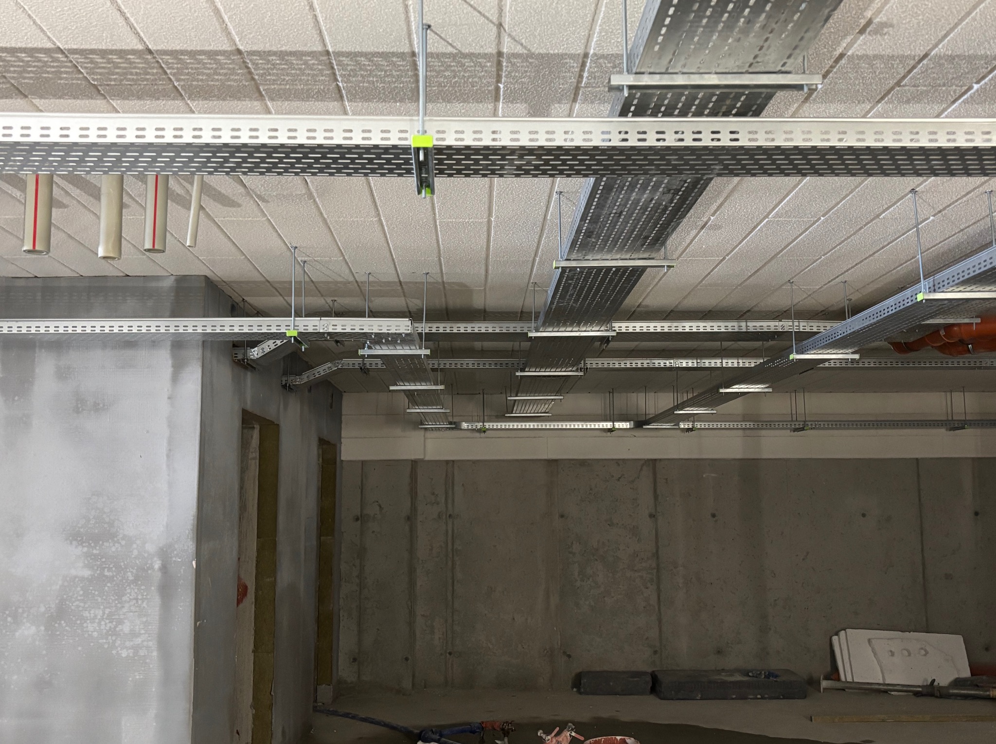 Sufit z widocznymi korytkami kablowymi i rurami instalacyjnymi, surowy stan wykończenia wnętrza, betonowe ściany. Widok z dołu.