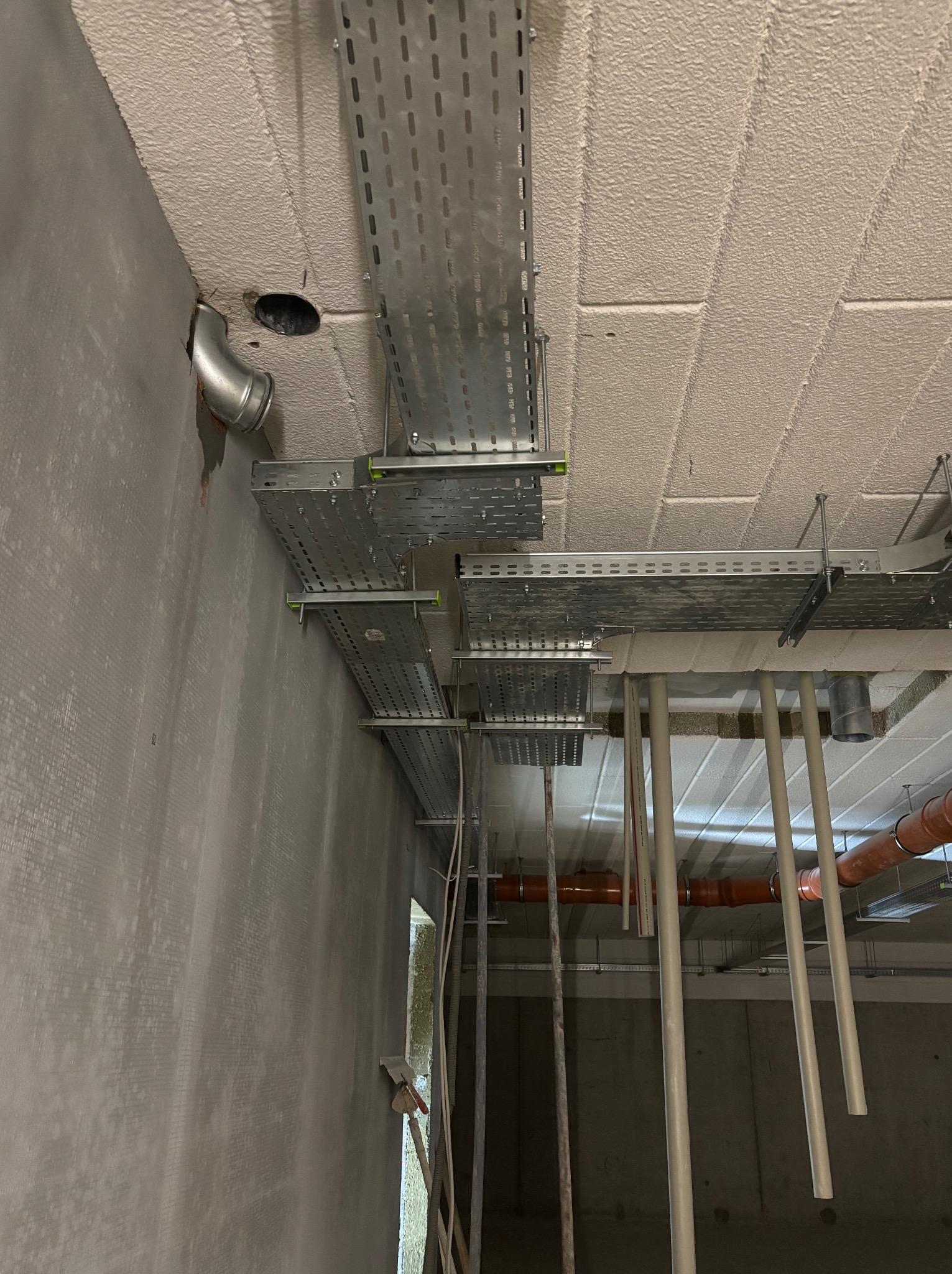 Montaż metalowych korytek kablowych pod sufitem w surowym wnętrzu. Widoczne rury i otwory w suficie. Prace instalacyjne w trakcie realizacji.