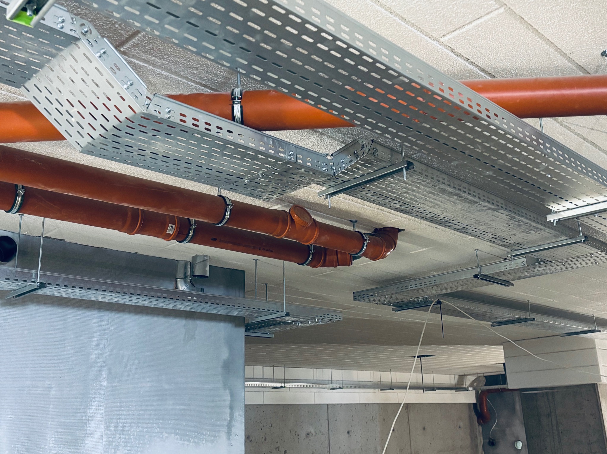 Montaż korytek kablowych i rur wentylacyjnych pod sufitem. Widoczne metalowe korytka z perforacją i pomarańczowe rury mocowane do sufitu za pomocą obejm.