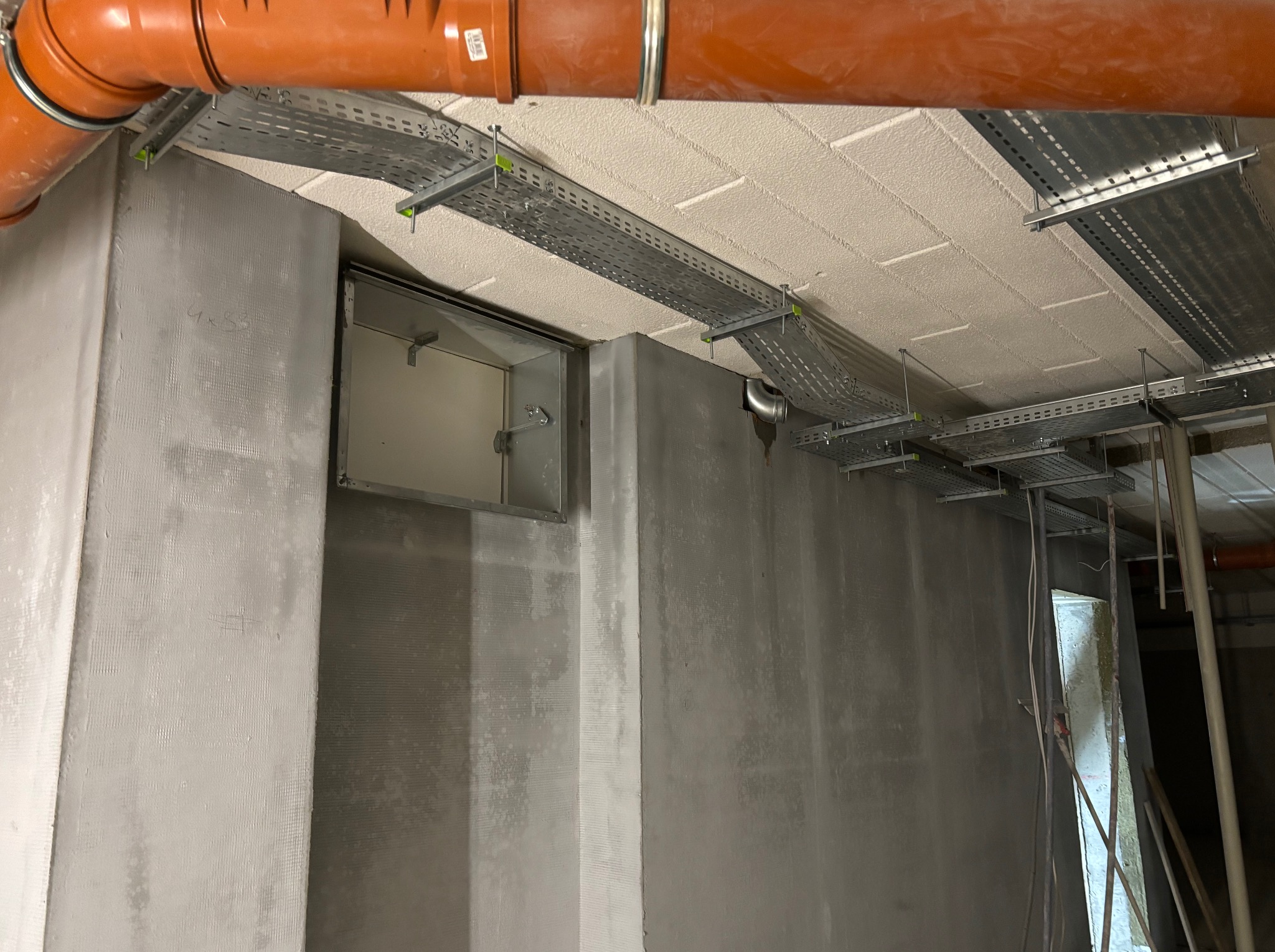 Szare ściany z zamontowanymi korytkami kablowymi pod sufitem i rurą. Widoczny otwór rewizyjny w ścianie. Surowy stan wykończenia wnętrza, instalacje w trakcie montażu.