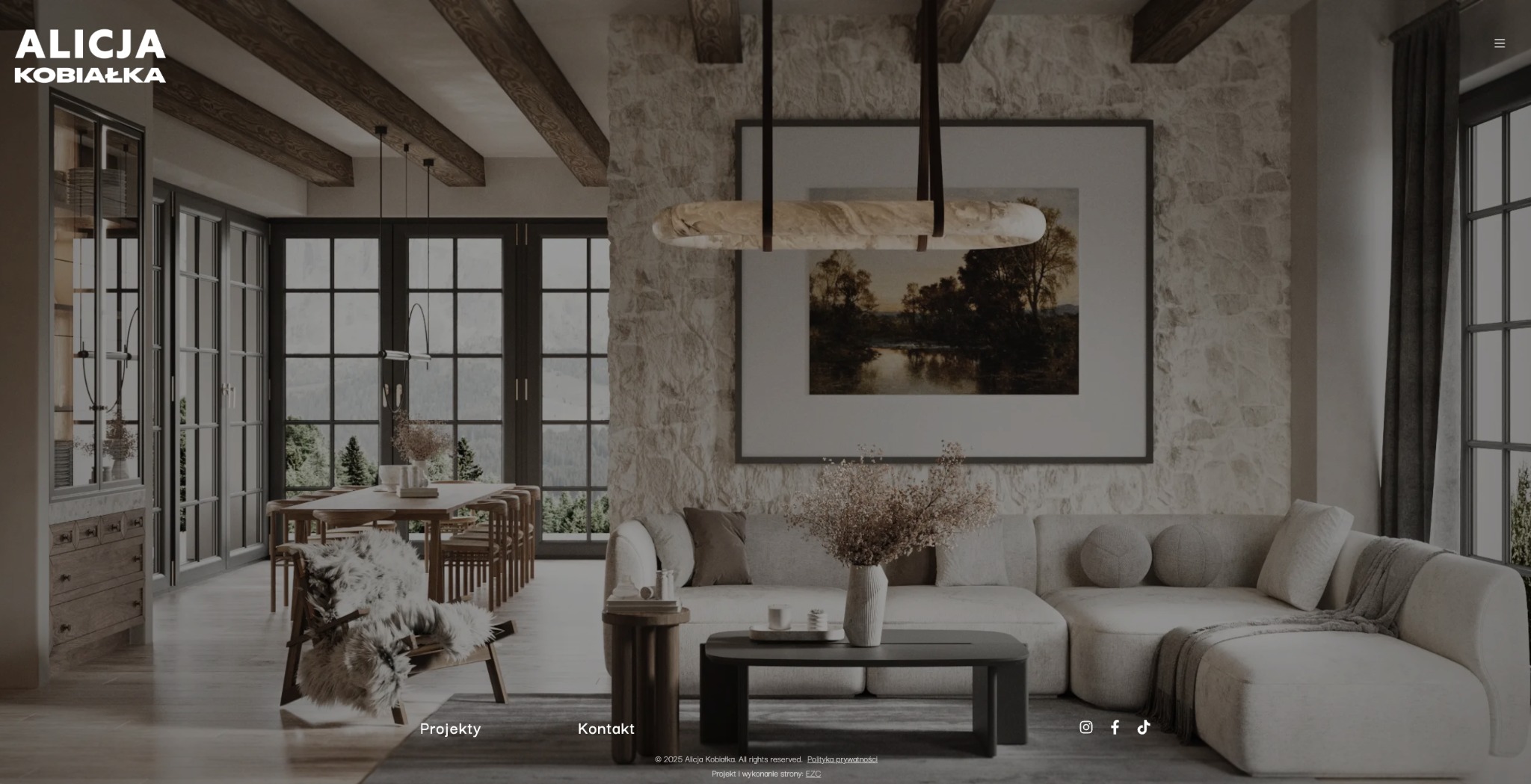 Strona www Alicji Kobiałki z widokiem na salon: sofa, stół, krzesła, okna. Minimalistyczny design, ciepłe barwy, kamienna ściana, obraz i drewniane belki stropowe.