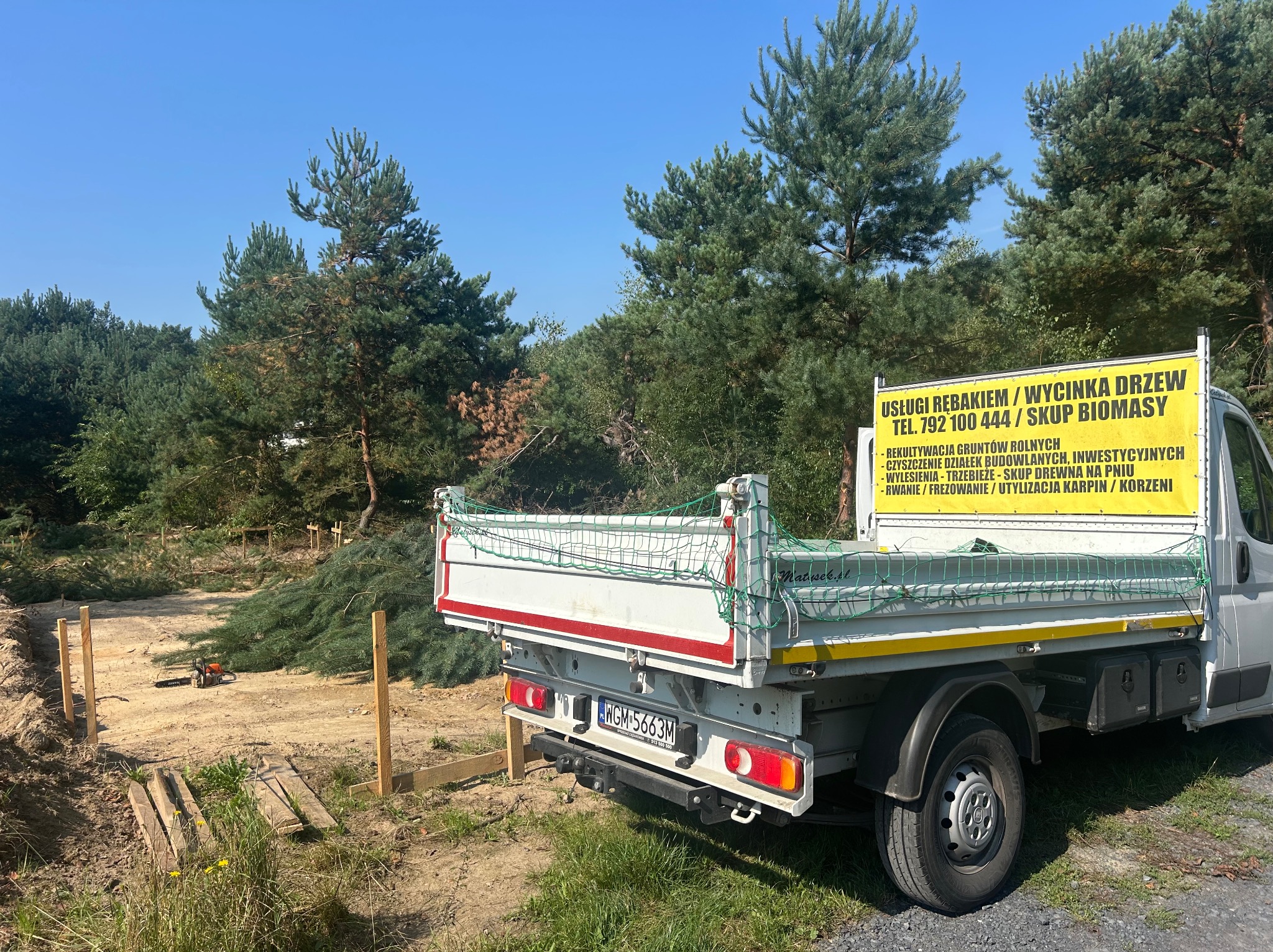 Ciężarówka z reklamą usług wycinki drzew i skupu biomasy w Sulechowie. W tle teren po wycince, piła łańcuchowa i ścięte drzewa.