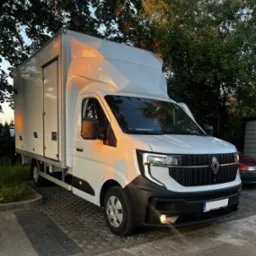 Biały samochód dostawczy Renault z wysoką zabudową, zaparkowany na brukowanej powierzchni, oświetlony popołudniowym słońcem, z widocznym logo Renault na grillu.