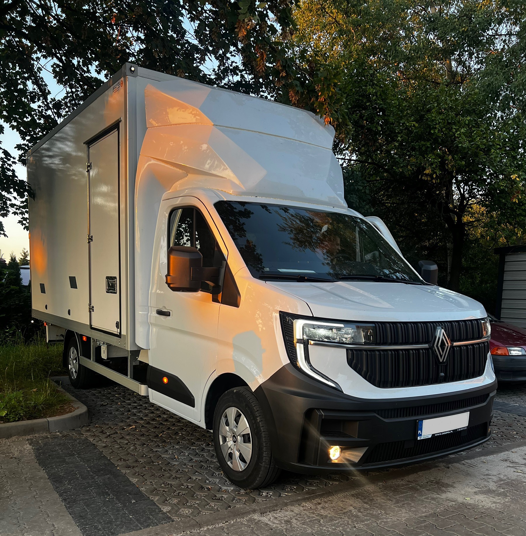 Biały samochód dostawczy Renault z wysoką zabudową, zaparkowany na brukowanej powierzchni, oświetlony popołudniowym słońcem, z widocznym logo Renault na grillu.