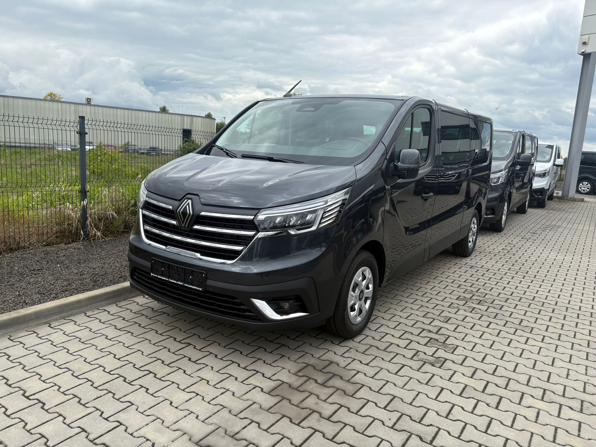 Szary Renault Trafic z chromowanymi detalami na kostce brukowej, ustawiony w rzędzie z innymi busami na tle pochmurnego nieba i metalowego ogrodzenia.