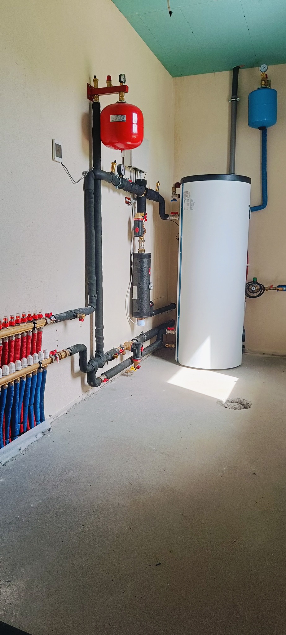 Instalacja CO z rozdzielaczem podłogowym, zbiornikiem buforowym i naczyniami przeponowymi w kotłowni. Rury w izolacji termicznej. Ściany w jasnym odcieniu beżu.