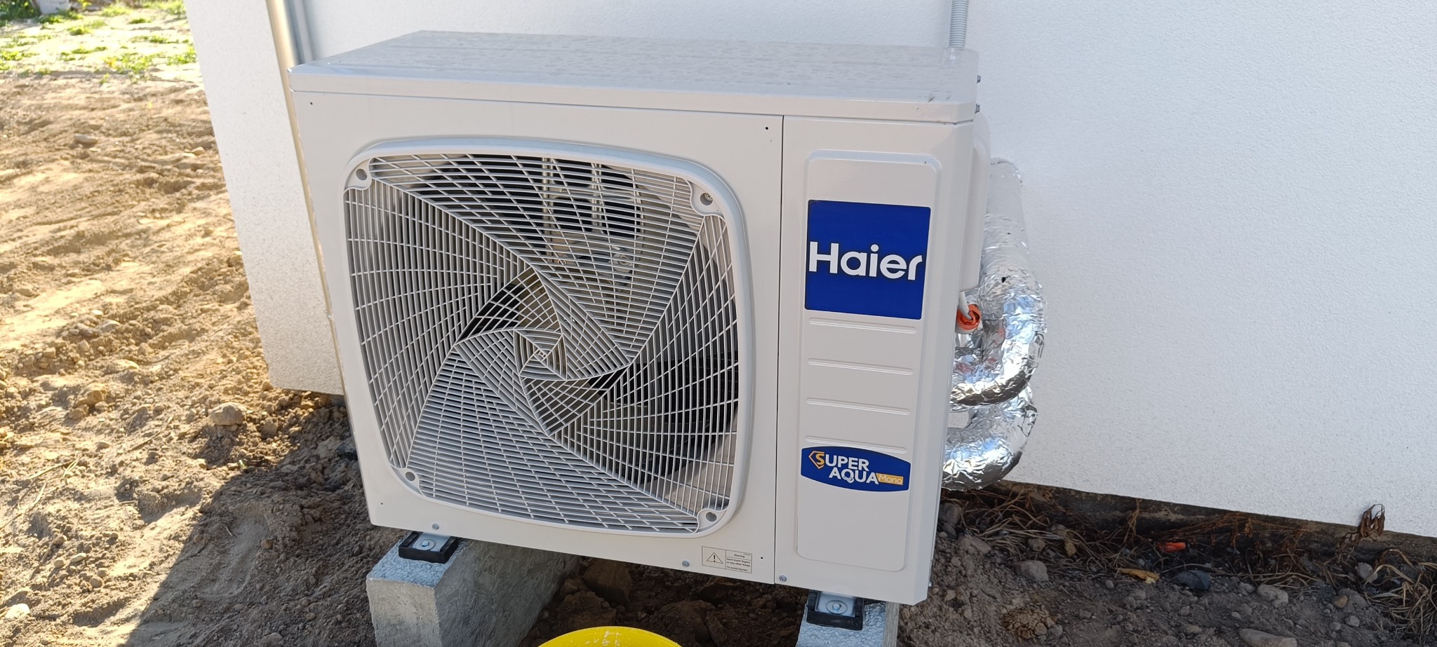 Biała, zewnętrzna jednostka klimatyzacji Haier Super Aqua Mono, zamontowana na betonowych podstawach przy ścianie budynku. Rury owinięte folią izolacyjną.