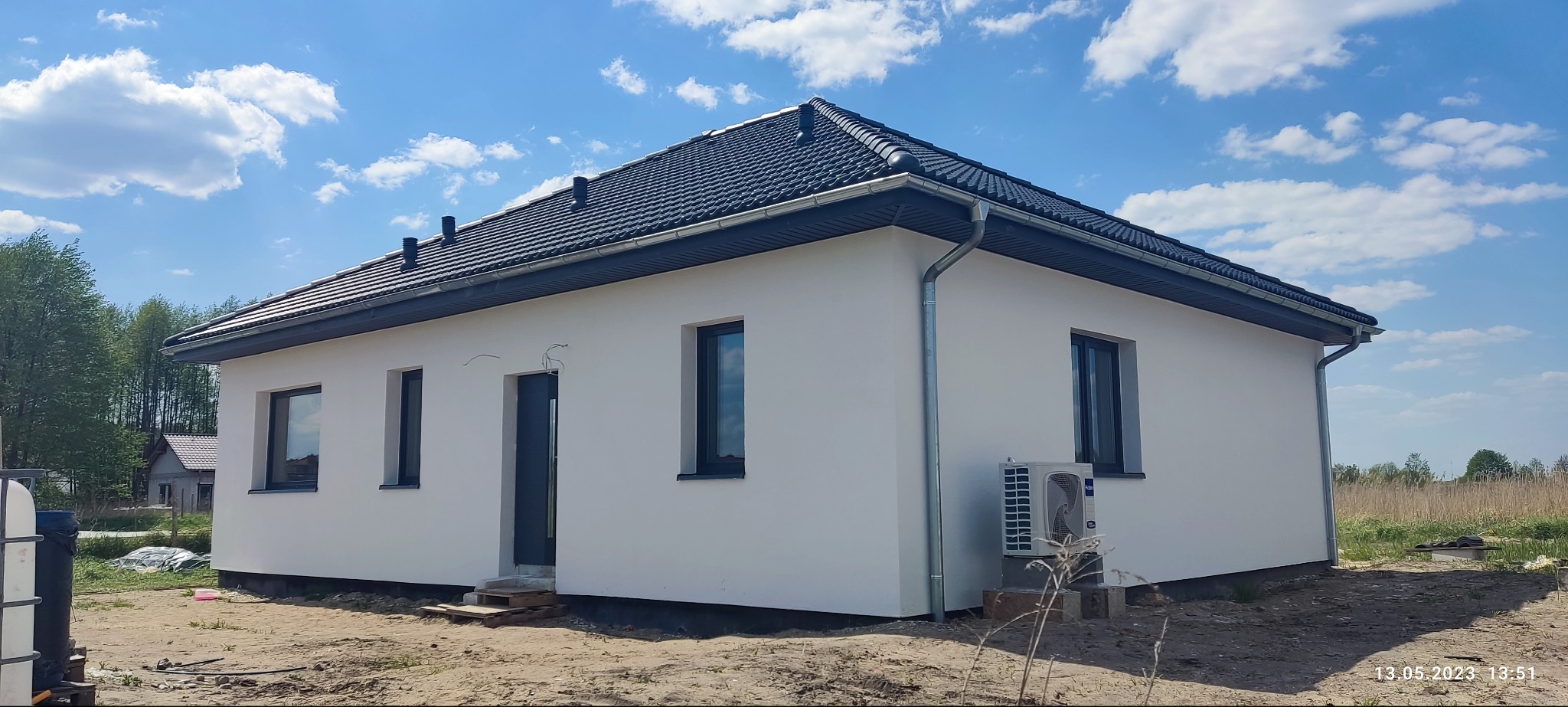 Nowoczesny, parterowy dom z klimatyzatorem na zewnątrz. Biała elewacja, ciemna dachówka i okna. Błękitne niebo z chmurami w tle. Widoczne rynny i fragment ogrodu.