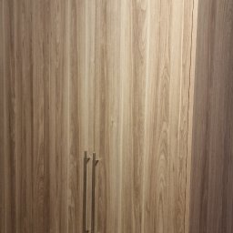 Krzysztof Zdunek usługi remontowe budowlane - Drewniana szafa na wymiar z podłużnymi uchwytami, dopasowana do skosu sufitu, z widoczną strukturą drewna i subtelnym oświetleniem.