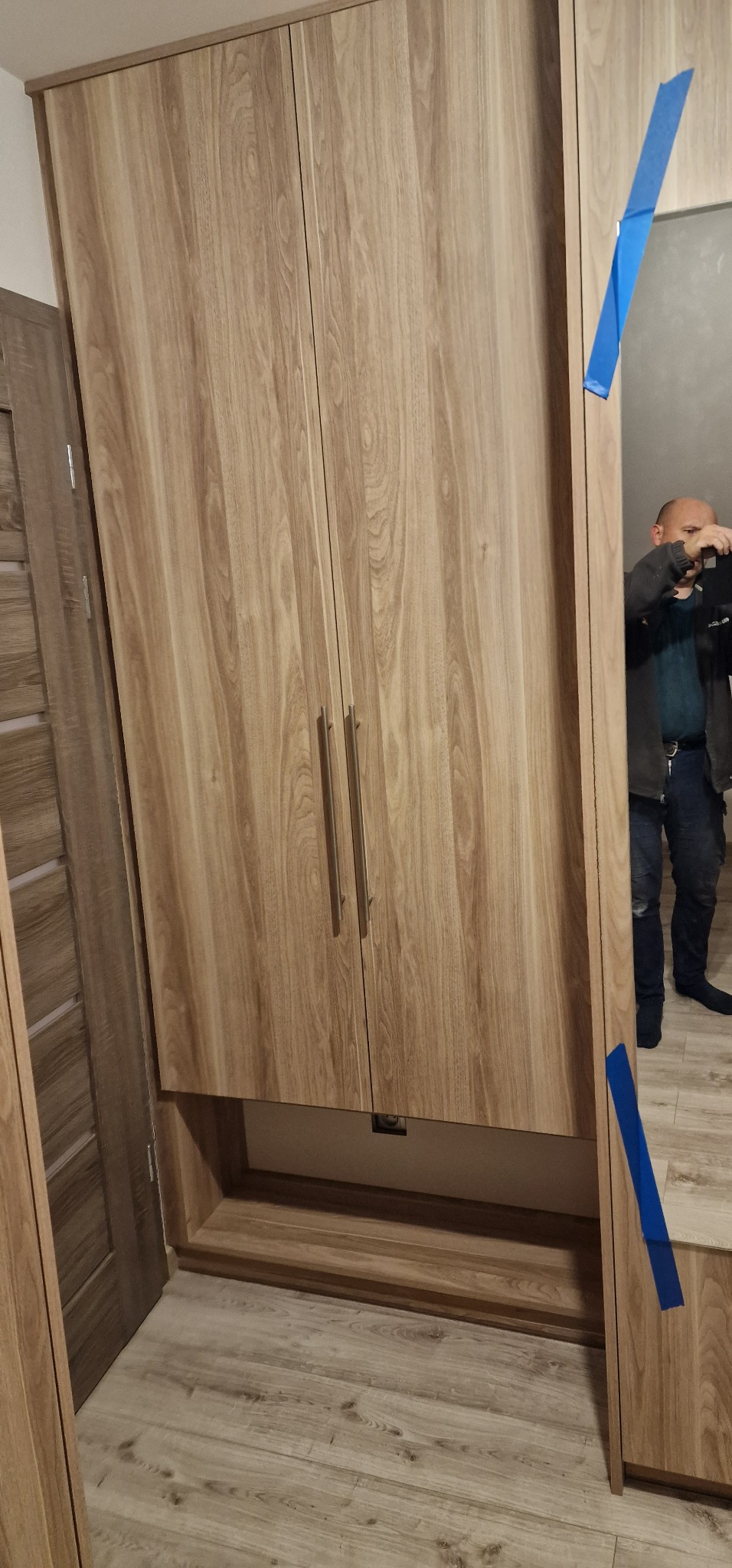 Szafa z drewnopodobnymi frontami, z długimi, pionowymi uchwytami. W lustrze odbija się osoba robiąca zdjęcie. Fragment drzwi po lewej stronie kadru.