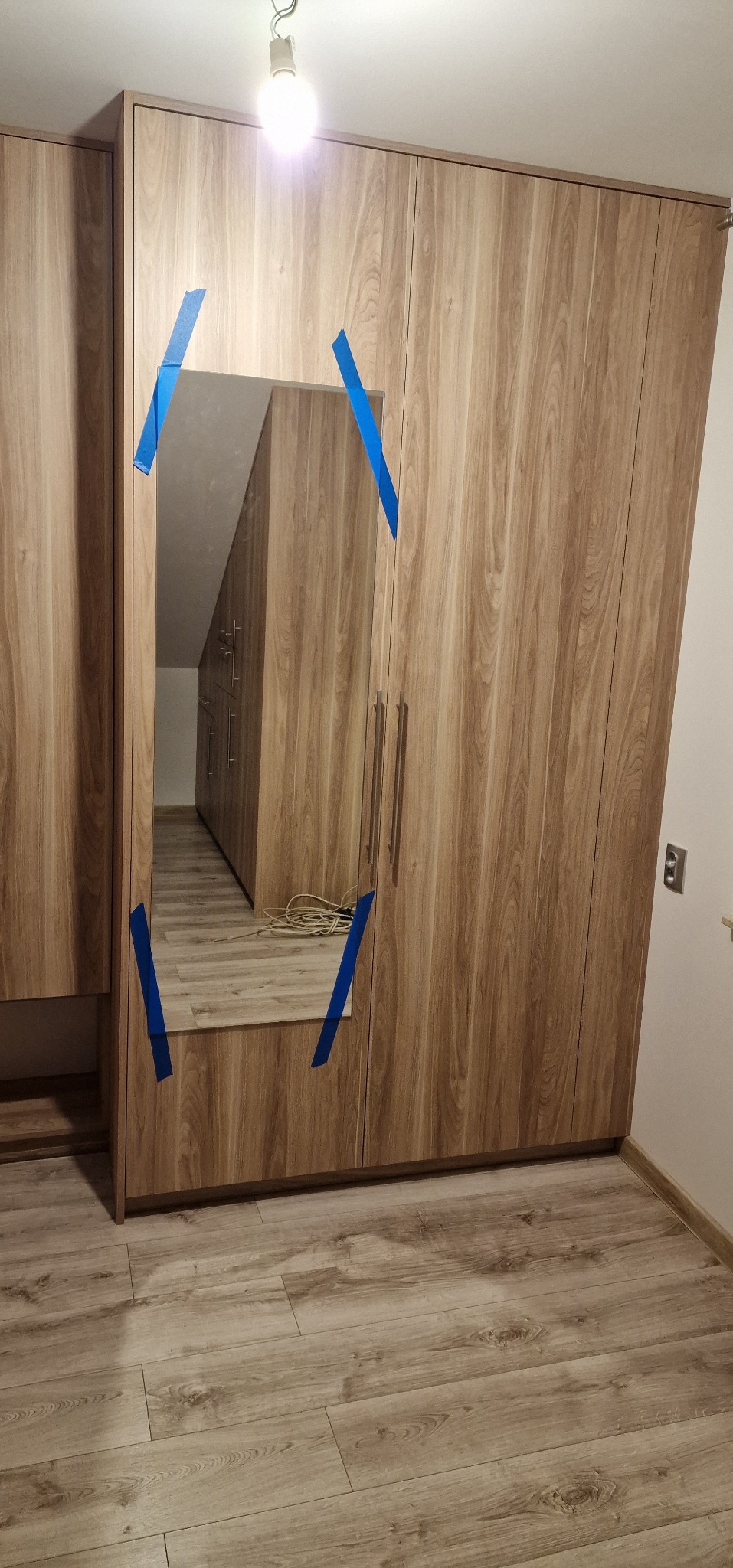 Szafa z lustrem, zabezpieczonym niebieską taśmą malarską. Widoczna podłoga z paneli. W lustrze odbicie wnętrza szafy.